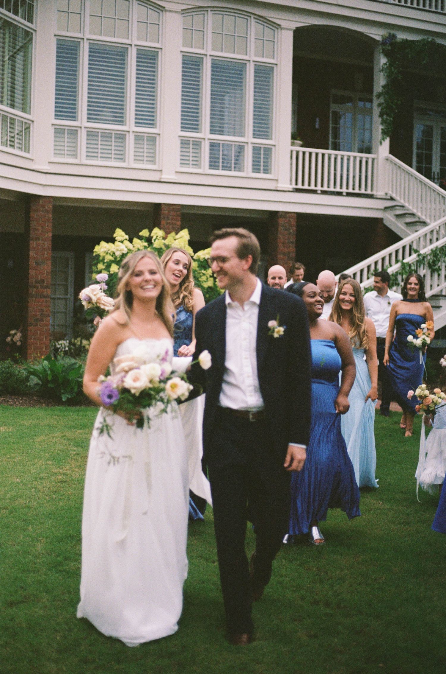 Smiling bride in white gown and groom in black suit walk through guests after outdoor wedding ceremony at estate venue.