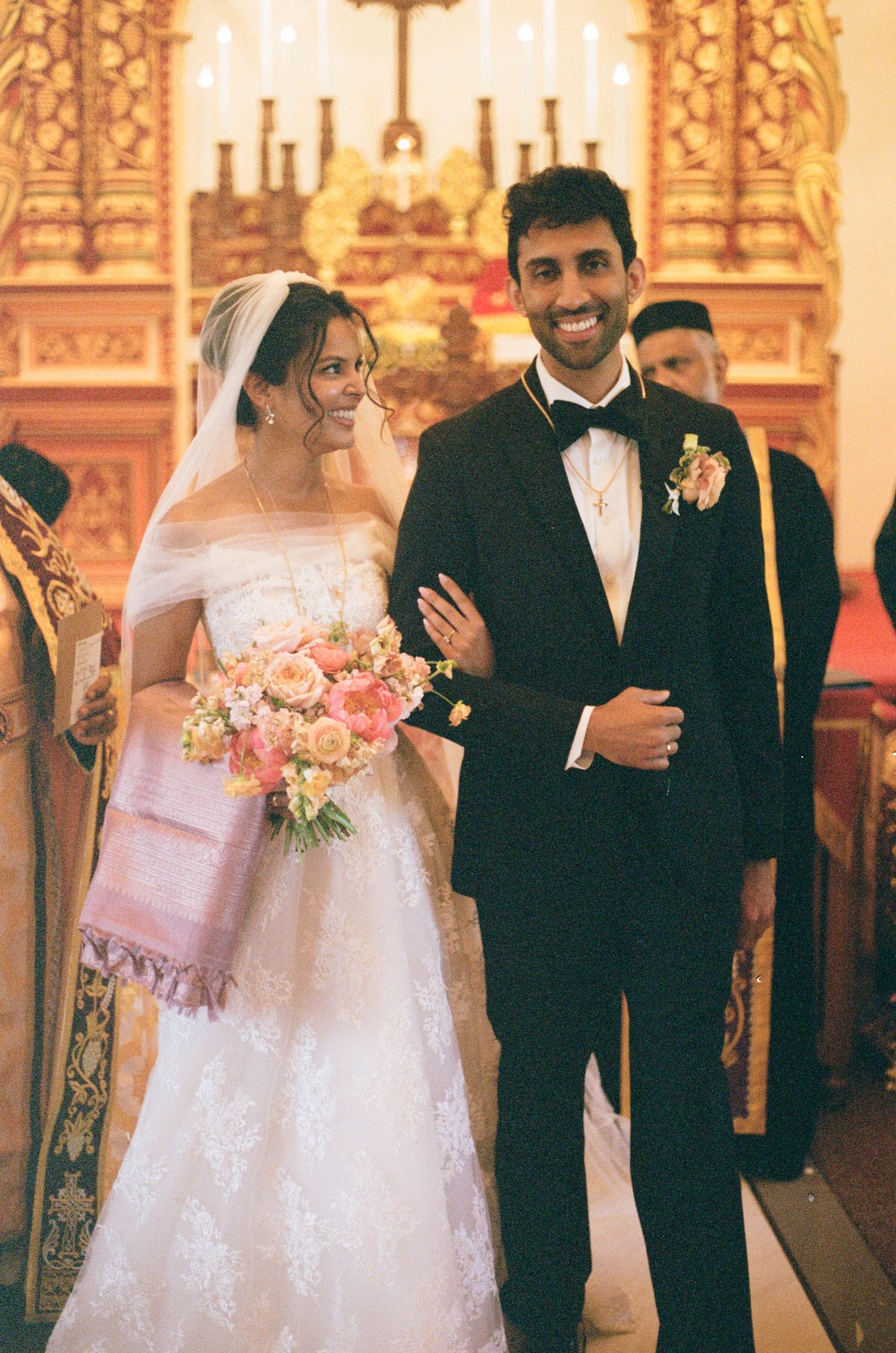 Smiling newlyweds walk down church aisle, bride in lace gown with pink bouquet, groom in black tuxedo.