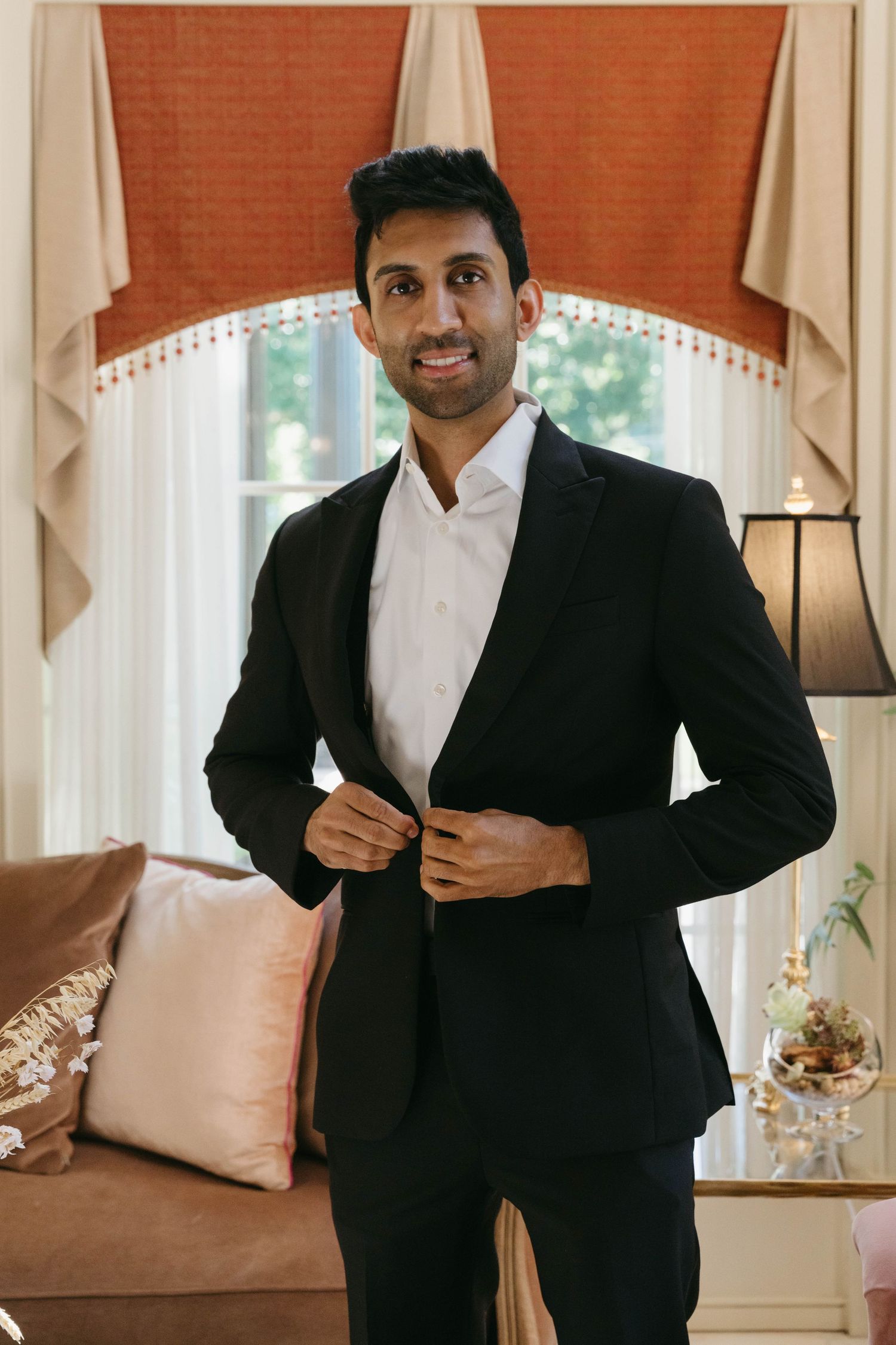 A well-dressed man in a black suit buttoning his jacket while standing in an elegant room with draped curtains.