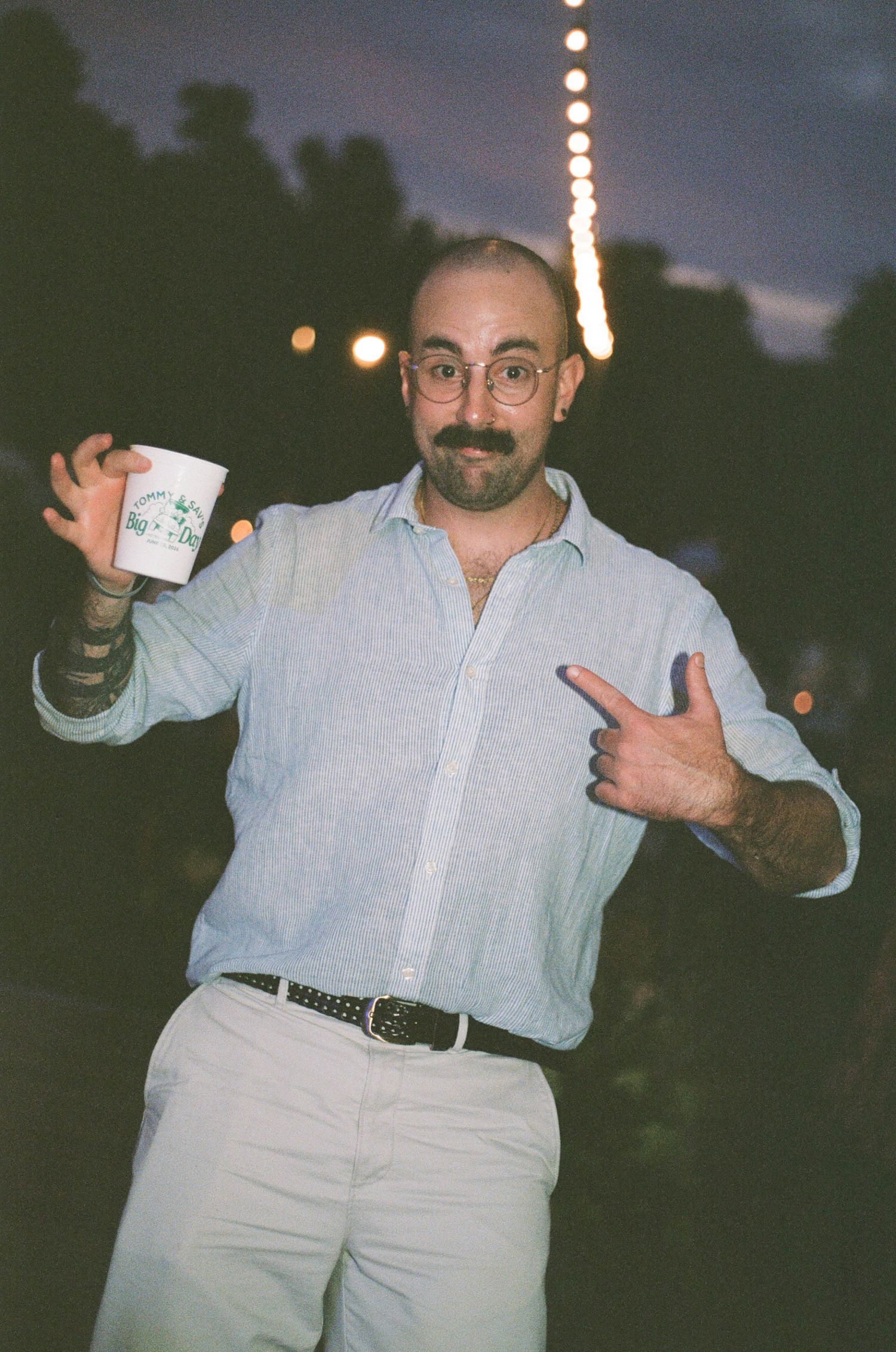 Smiling bald man with mustache points to a cup at an outdoor evening wedding event.