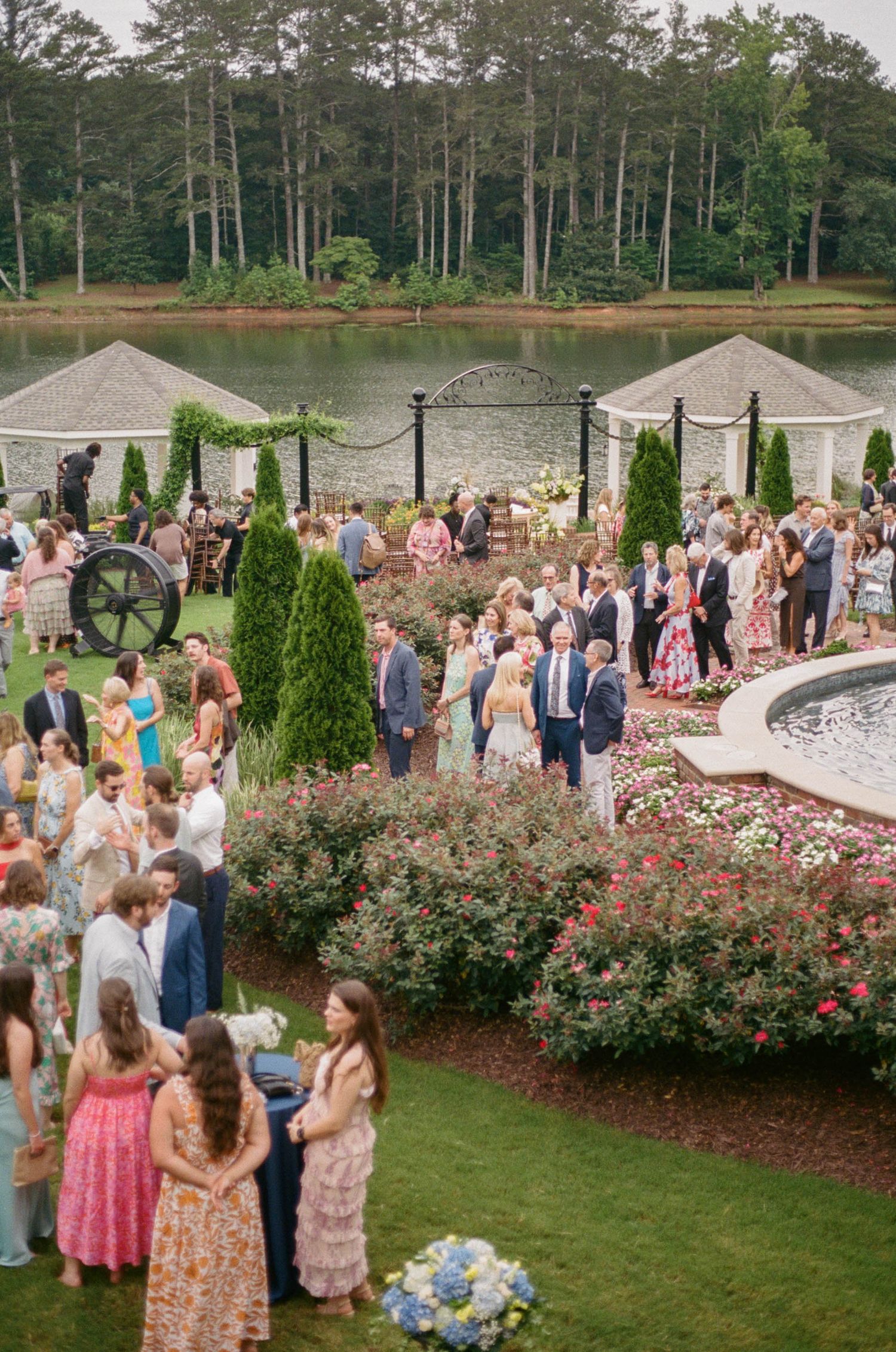 Aerial view of elegant garden wedding reception with guests mingling near fountain and lake backdrop.