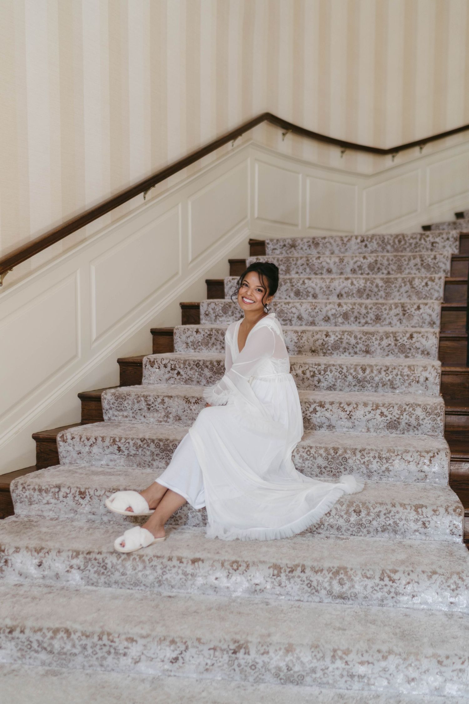 Smiling bride in white wedding gown and slippers sitting on marble staircase in luxurious venue.