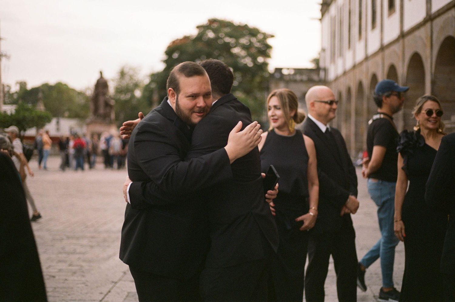 Guests in formal black attire mingle at an outdoor event venue with stone columns.