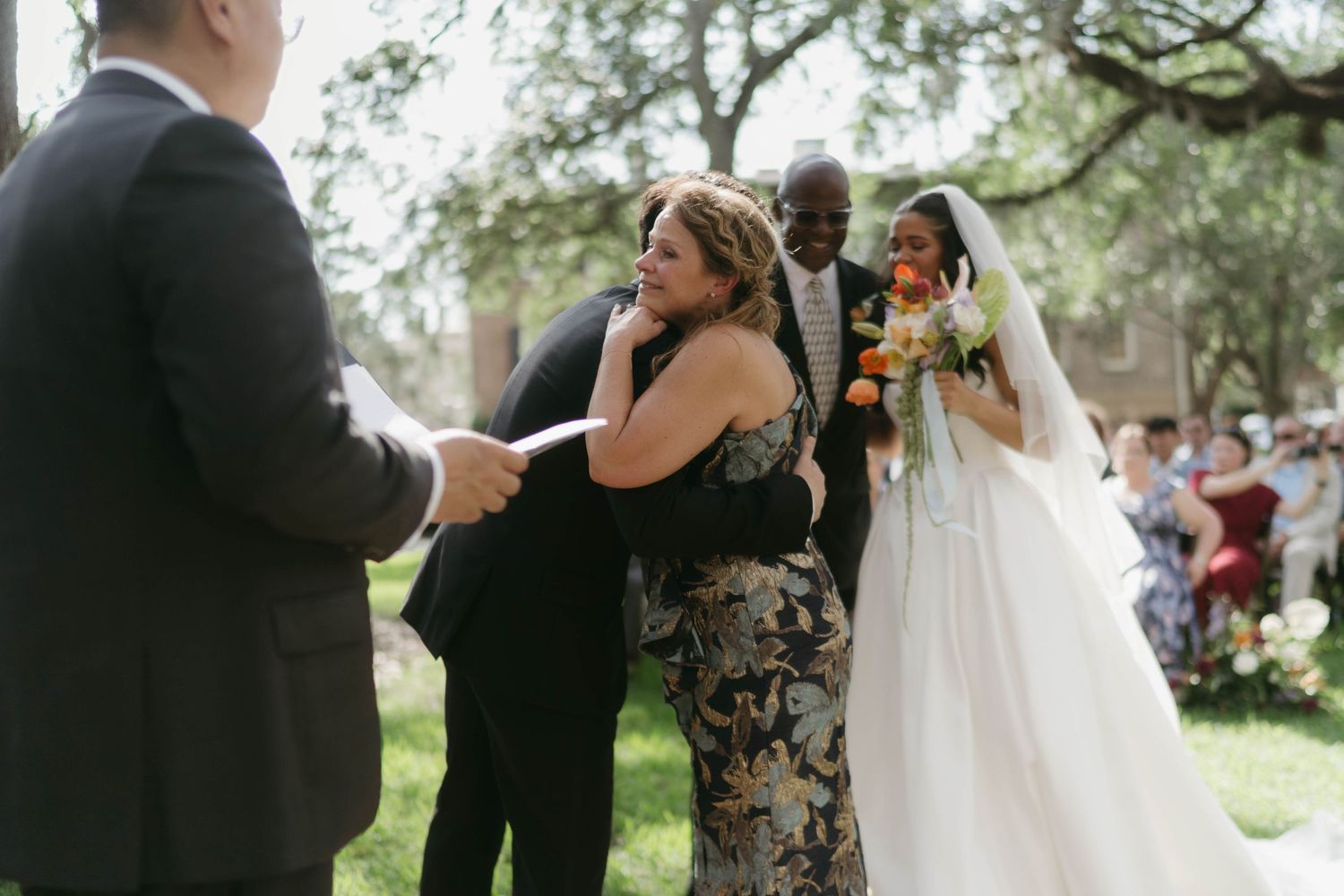 An outdoor wedding ceremony taking place under large oak trees.