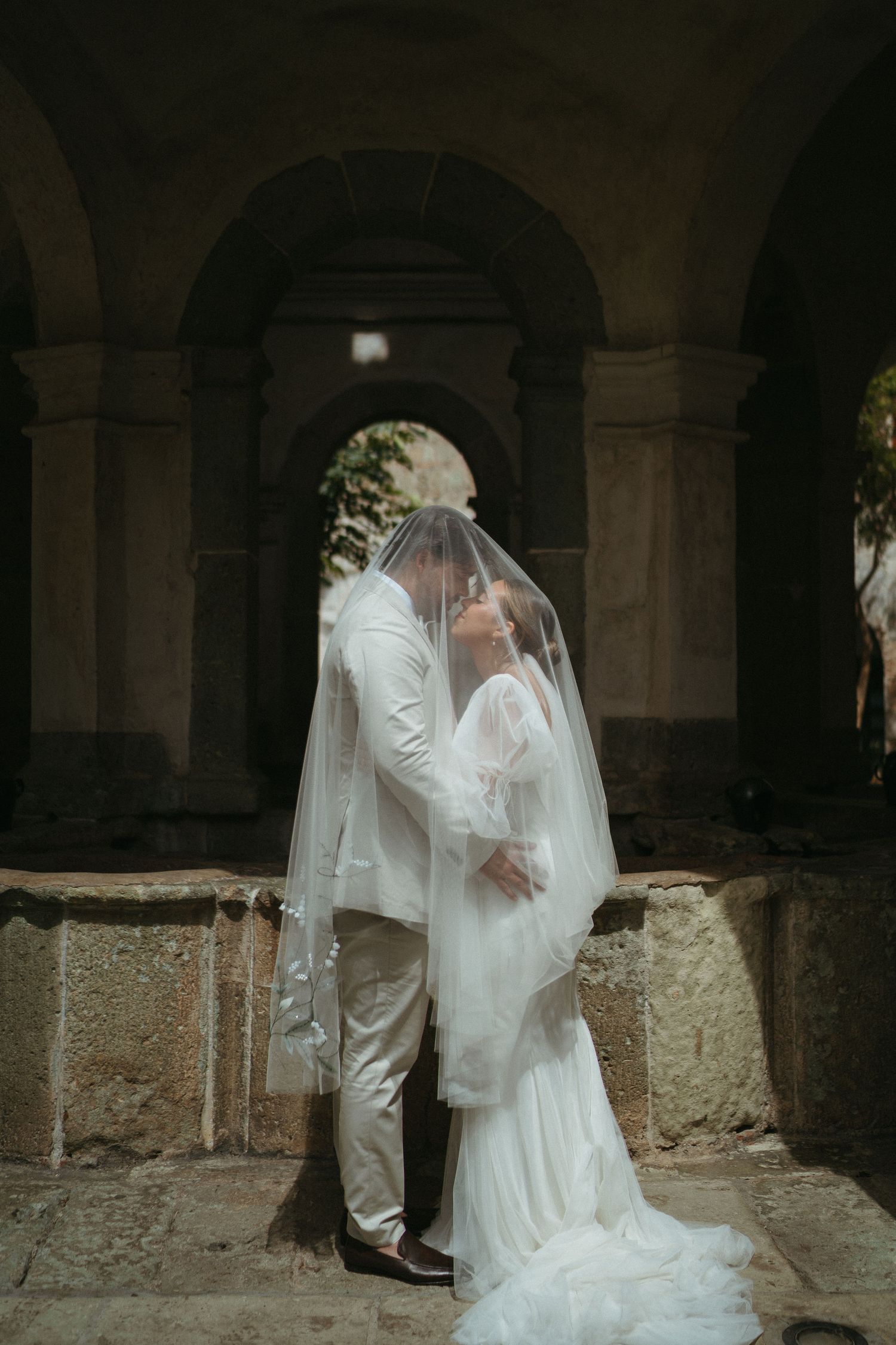 A romantic wedding pose under historic stone archways with flowing white dress and veil.
