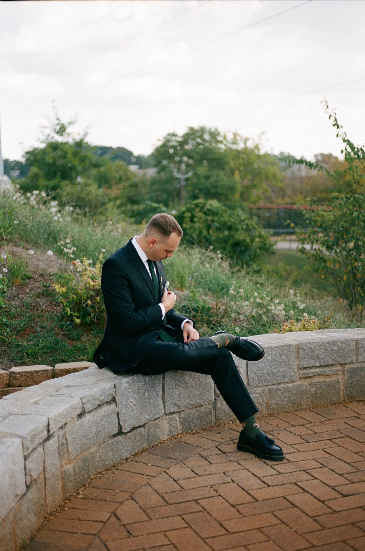 A person in a dark suit sits contemplatively on curved brick steps surrounded by lush greenery.