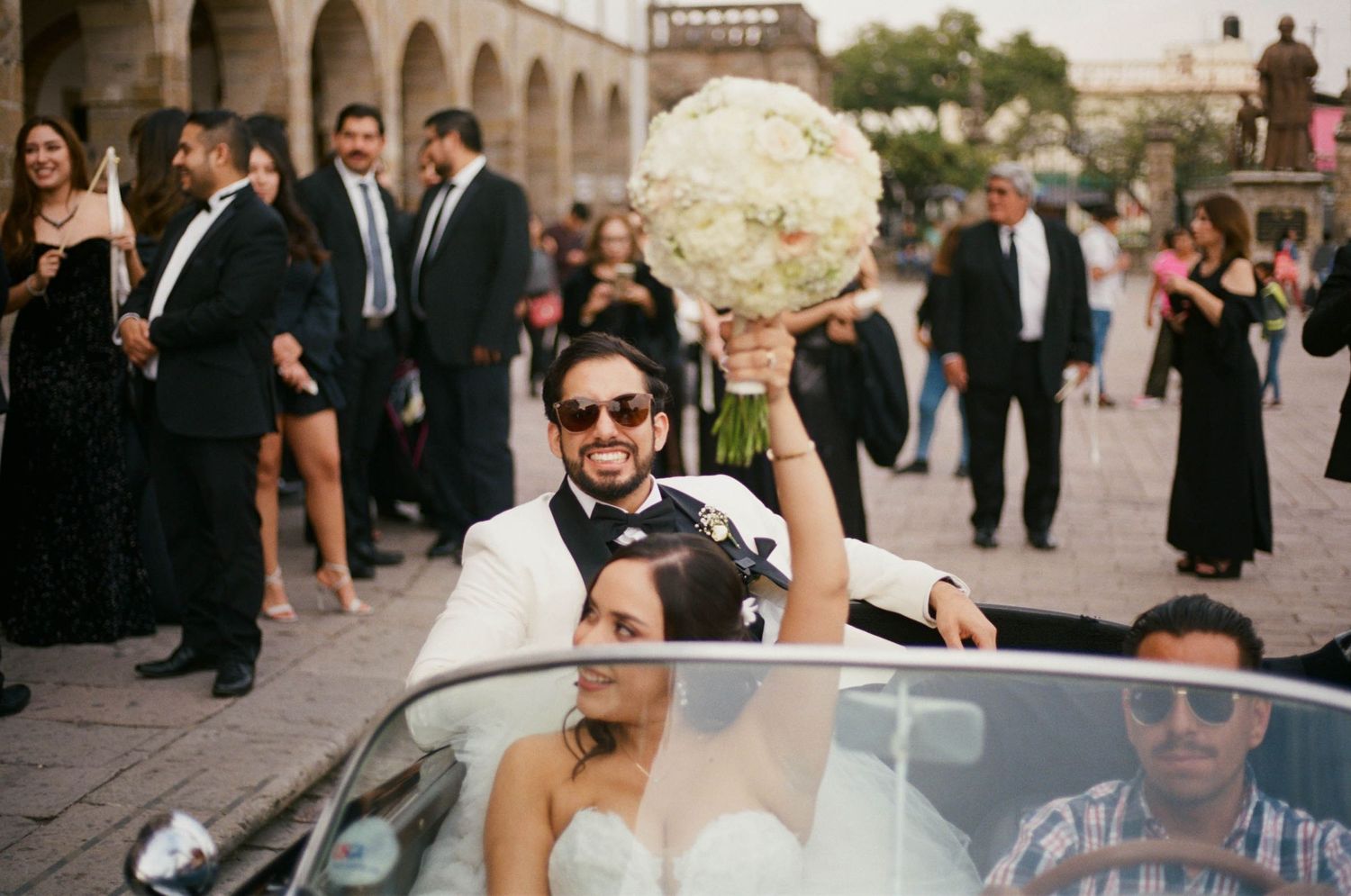 Wedding party gathers around a classic black convertible car outside a historic venue.