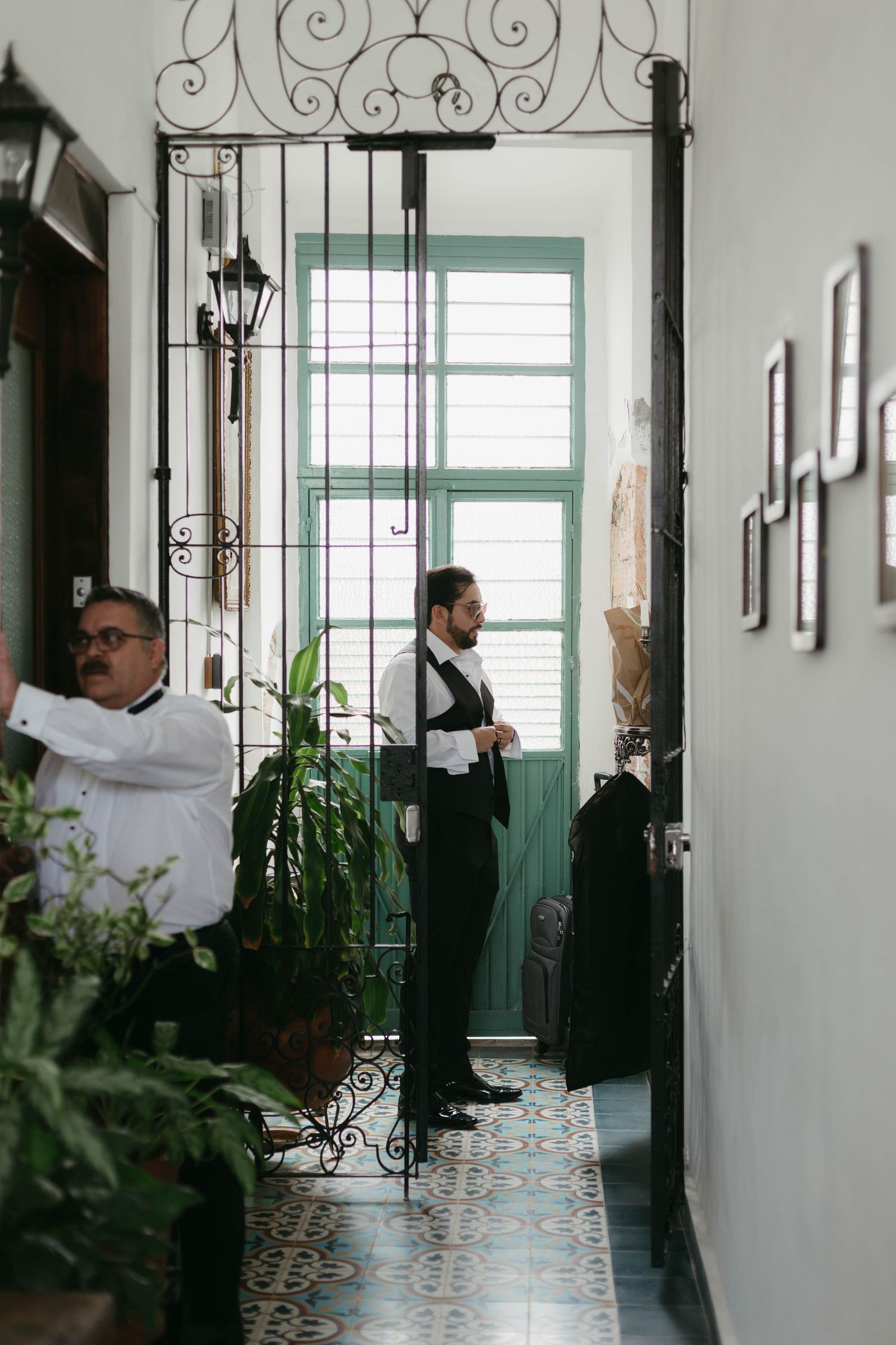 People in white clothing walk through an elegant corridor with wrought iron details and tiled floors.