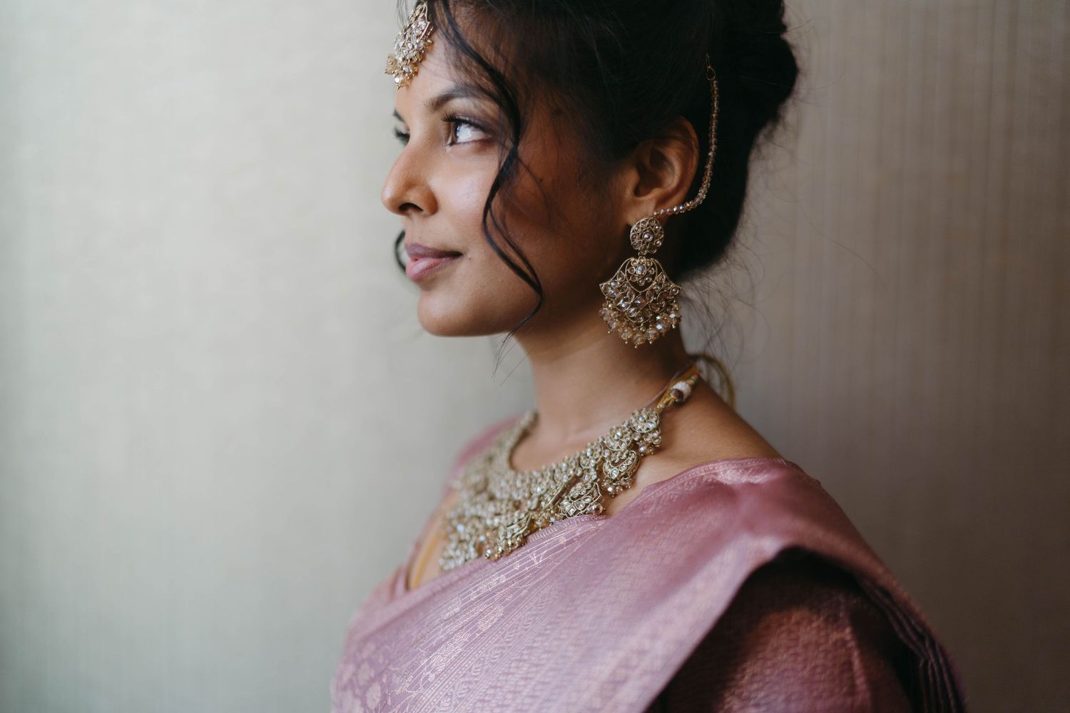 Indian bride in pink saree wearing ornate gold jewelry and maang tikka poses gracefully for bridal portrait.