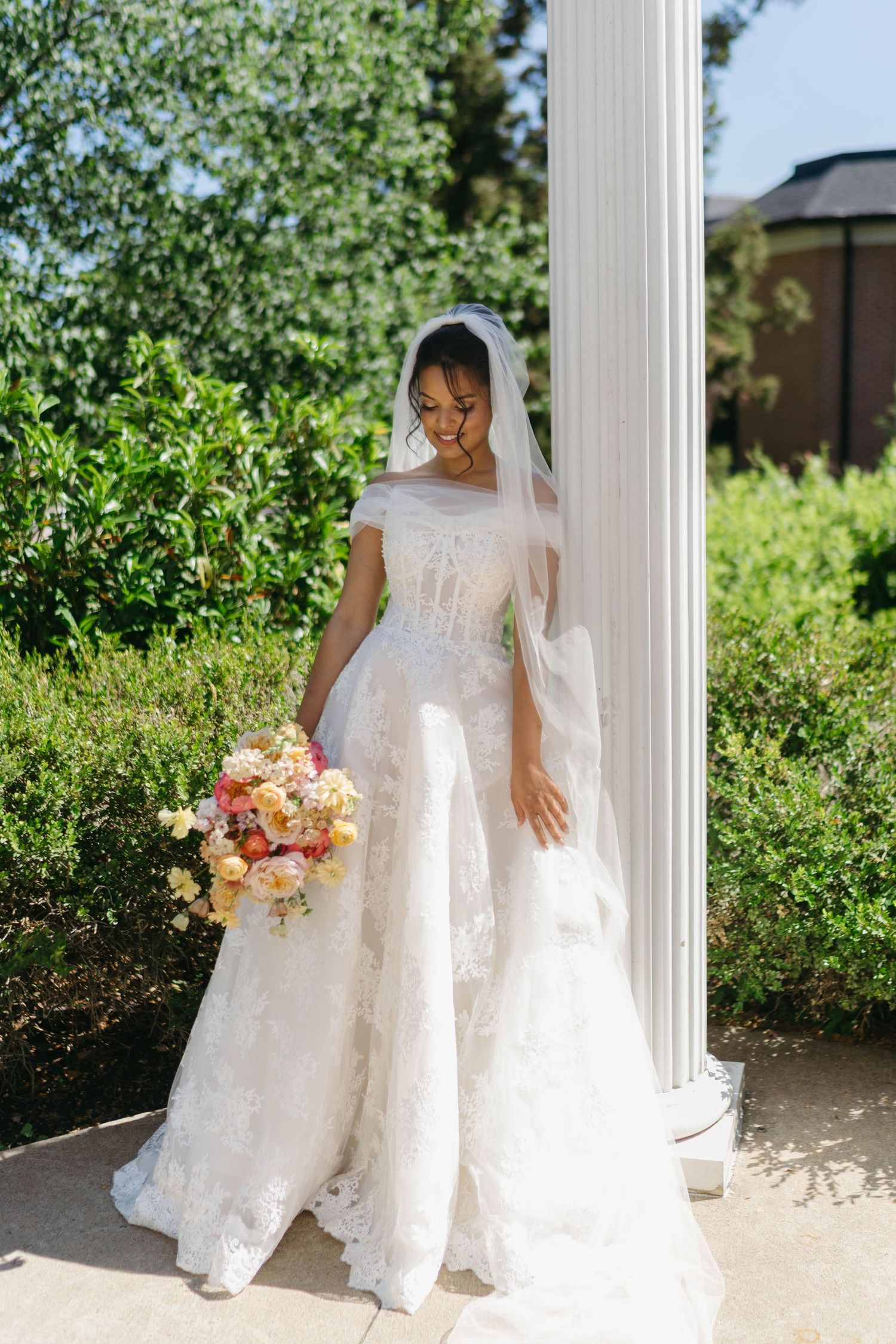 Smiling bride in off-shoulder lace wedding dress holding colorful bouquet beside white column outdoors.