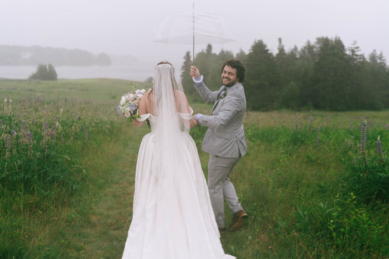 A couple in wedding attire walks through a green meadow on an overcast day.