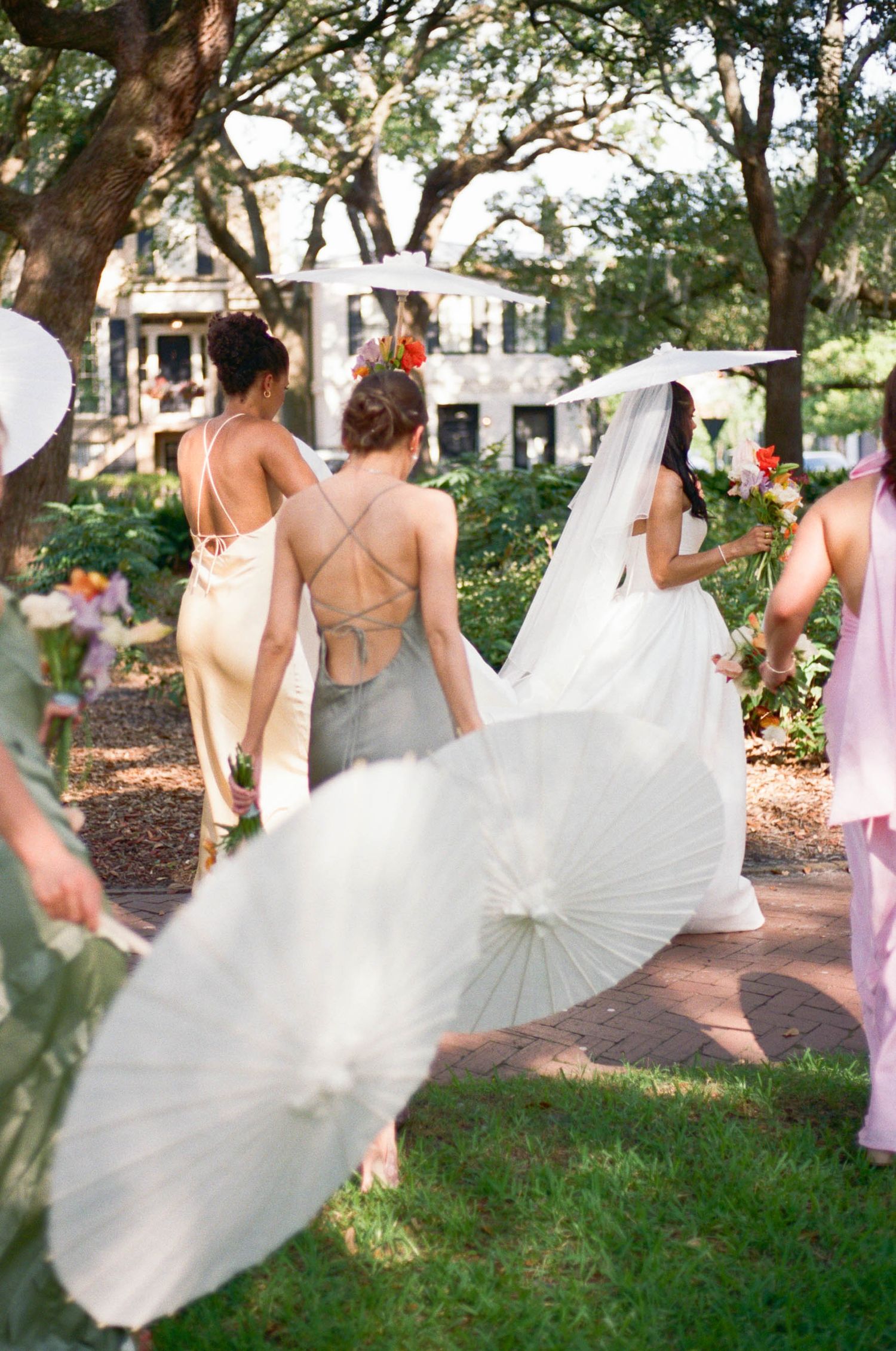Bridesmaids in light dresses carry parasols while walking along a garden path.