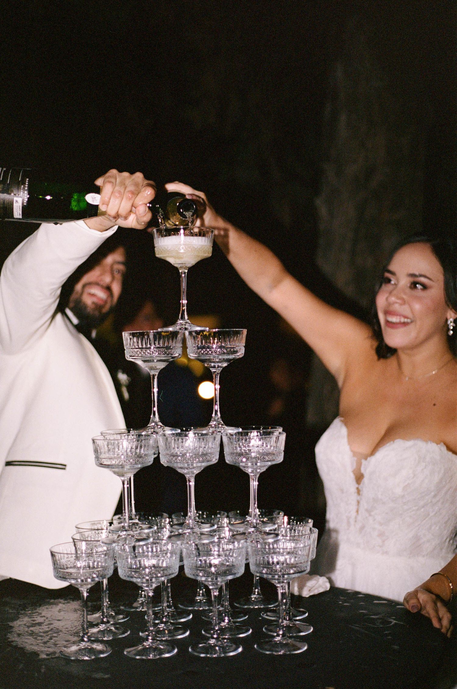 Wedding couple pouring champagne into a multi-tiered glass tower during their reception celebration.