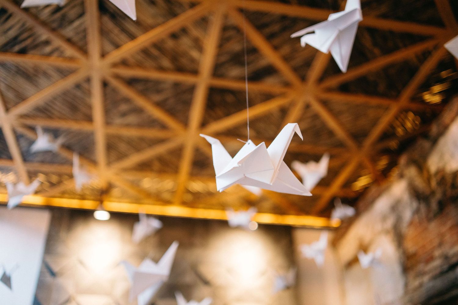 Wedding guests celebrate under a rustic wooden ceiling decorated with white paper cranes and string lights.