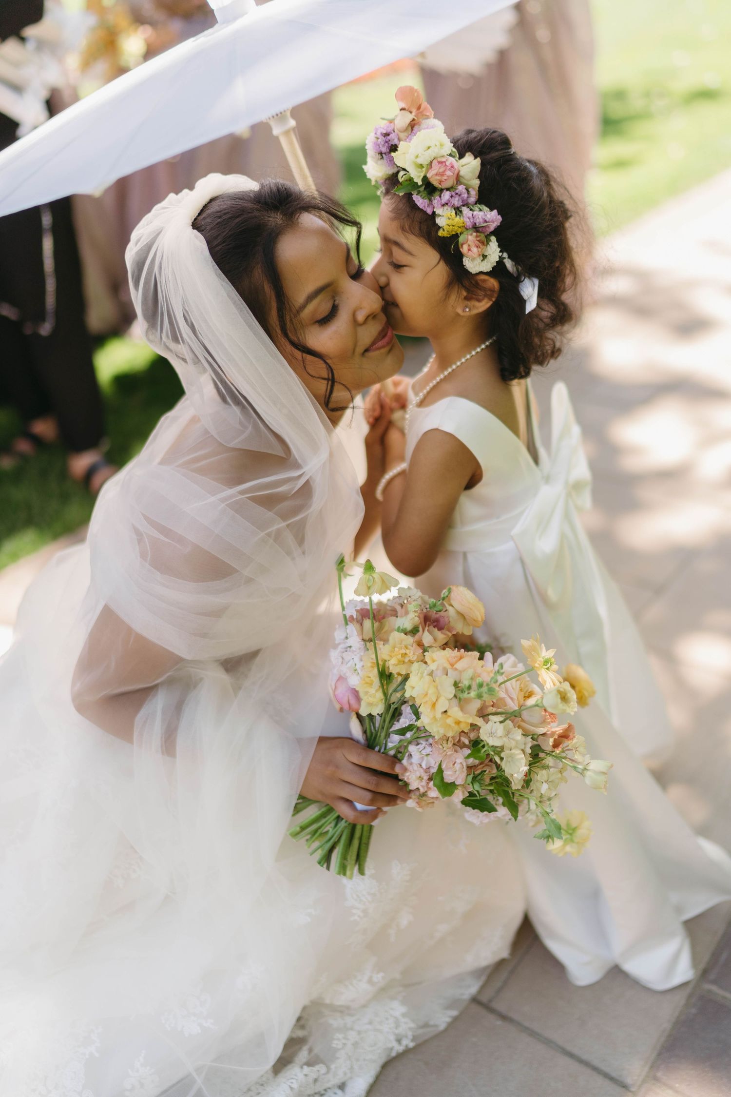 A bride in a white lace gown and veil holds a pastel bouquet while a flower girl in white kisses her cheek.