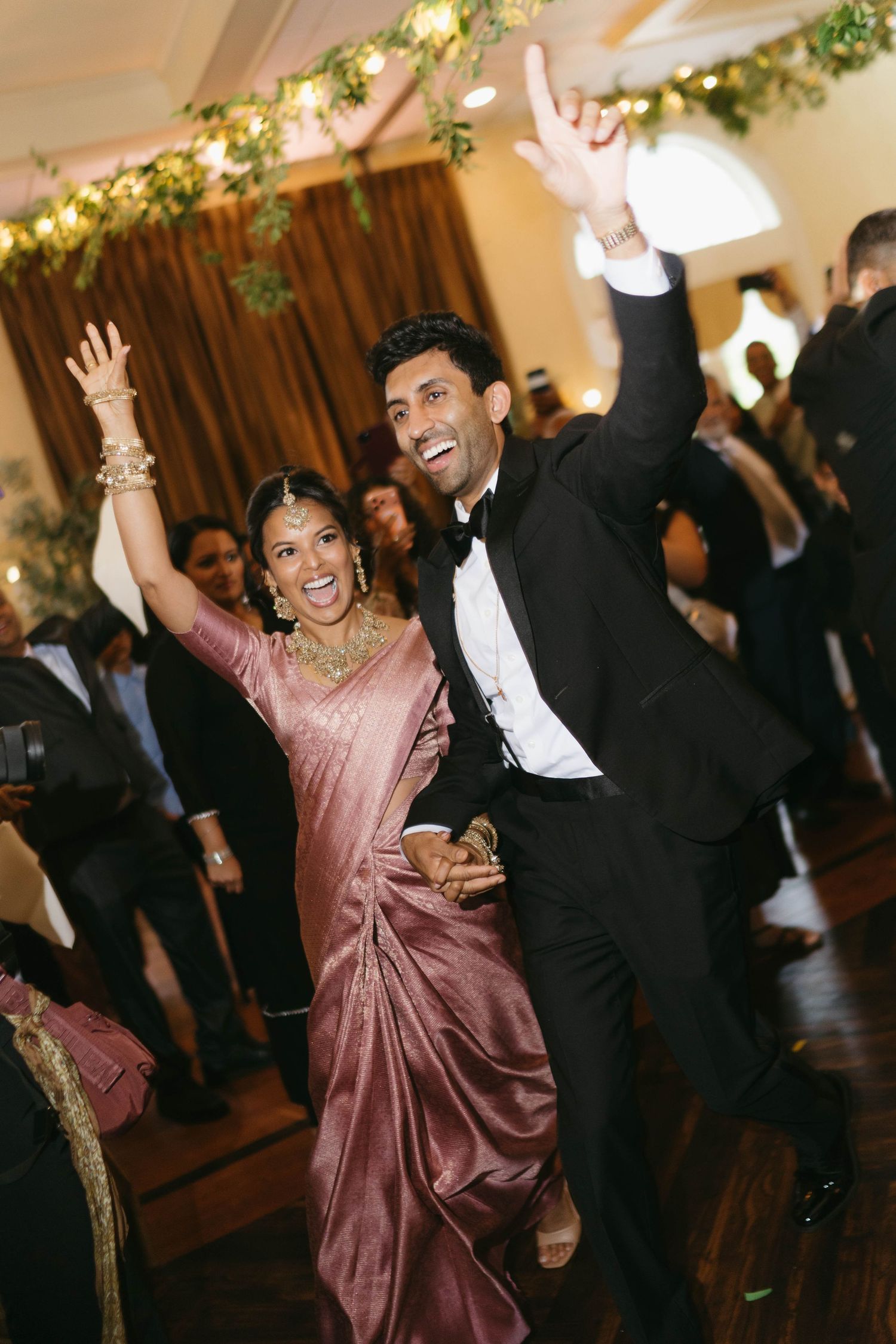 Joyful Indian couple celebrating at wedding reception, bride in pink saree with jewelry, groom in black tuxedo dancing.
