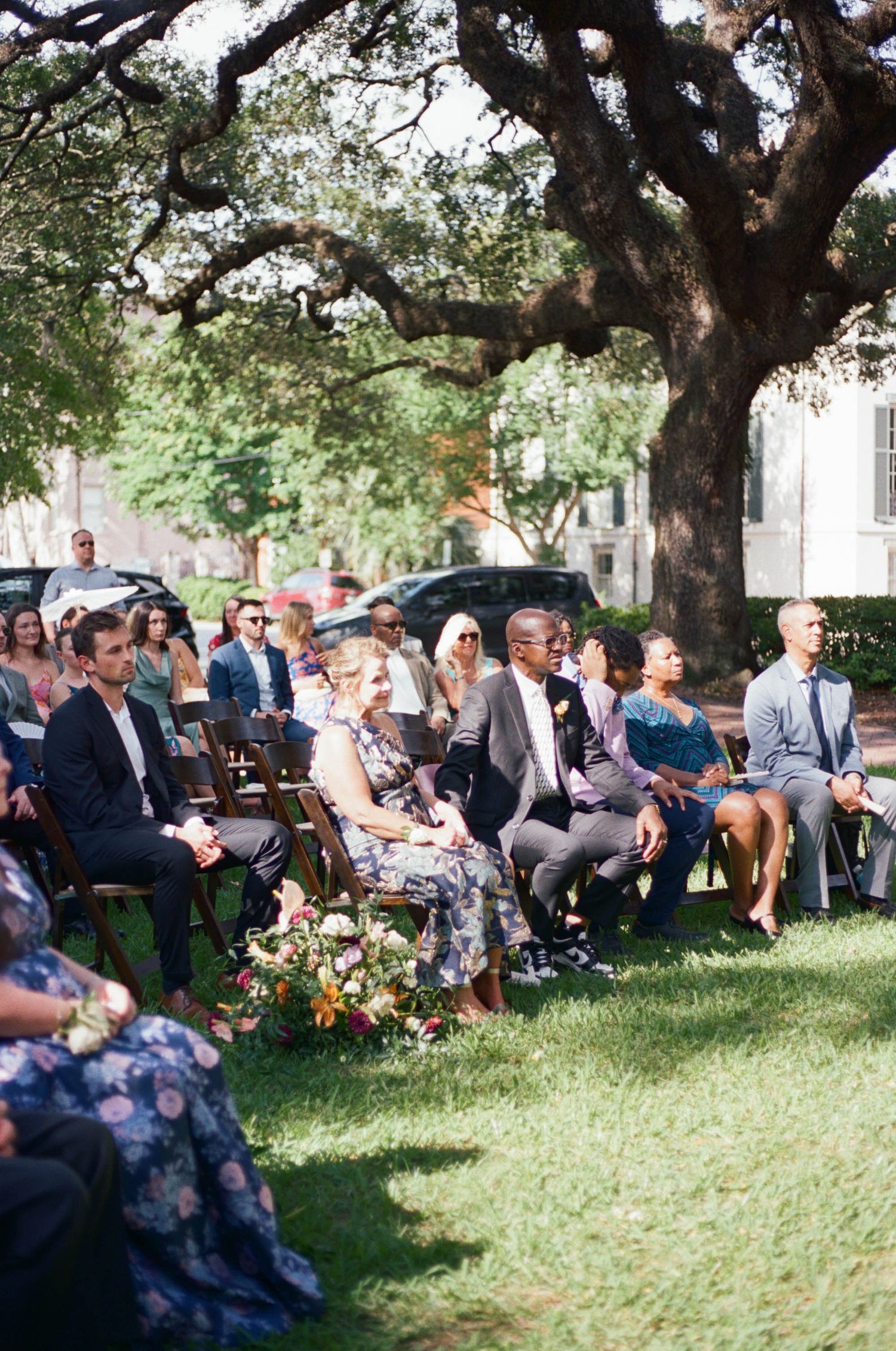 Wedding guests seated outdoors under a large oak tree for a ceremony.