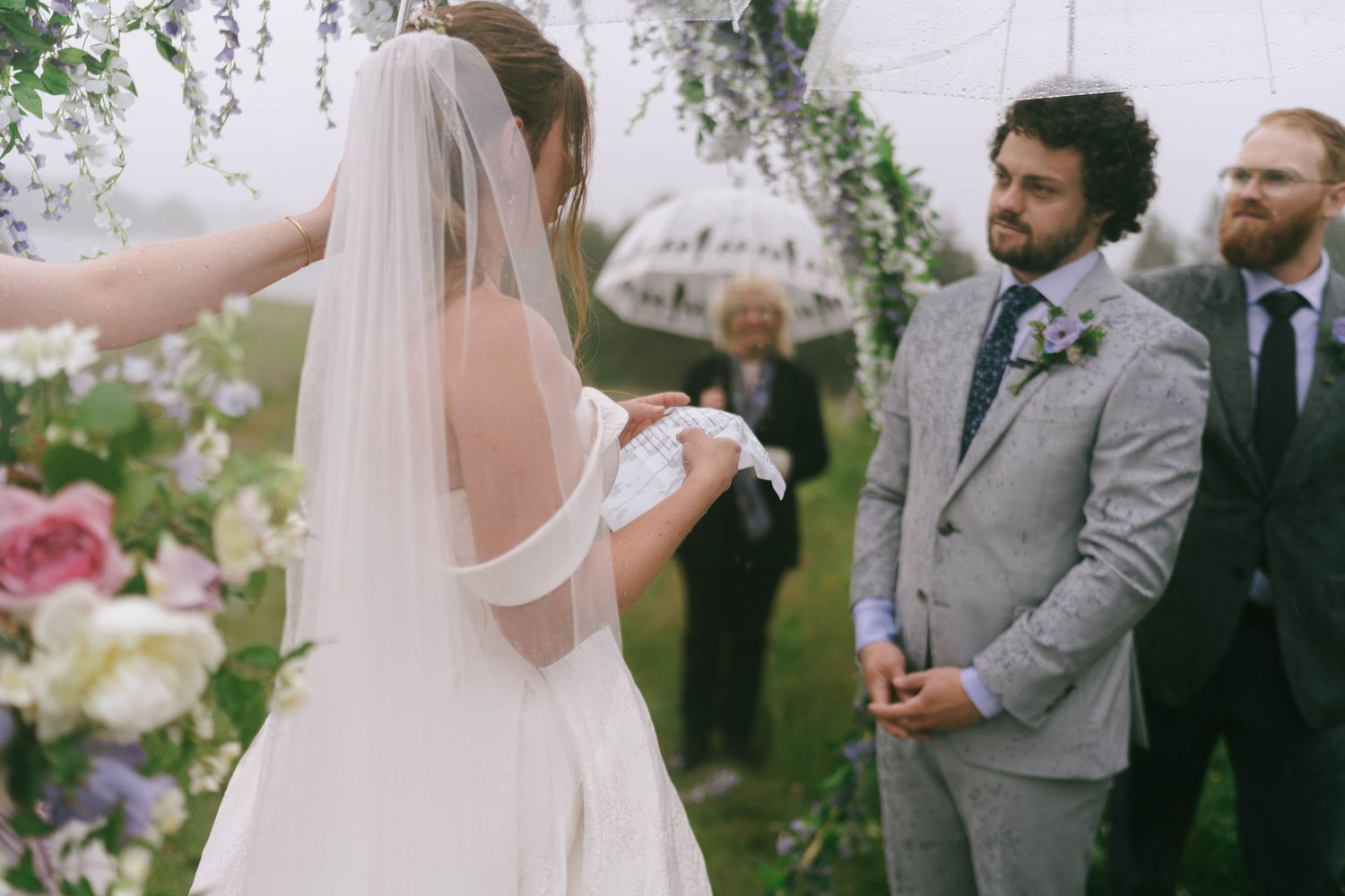 A wedding ceremony takes place under a floral arch decorated with pink roses and greenery.