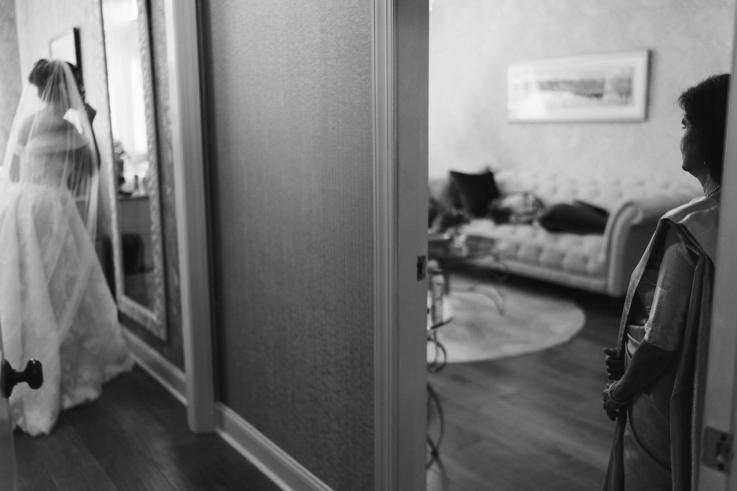 Black and white photo of bride in veil reflected in mirror while guest watches in hotel room.
