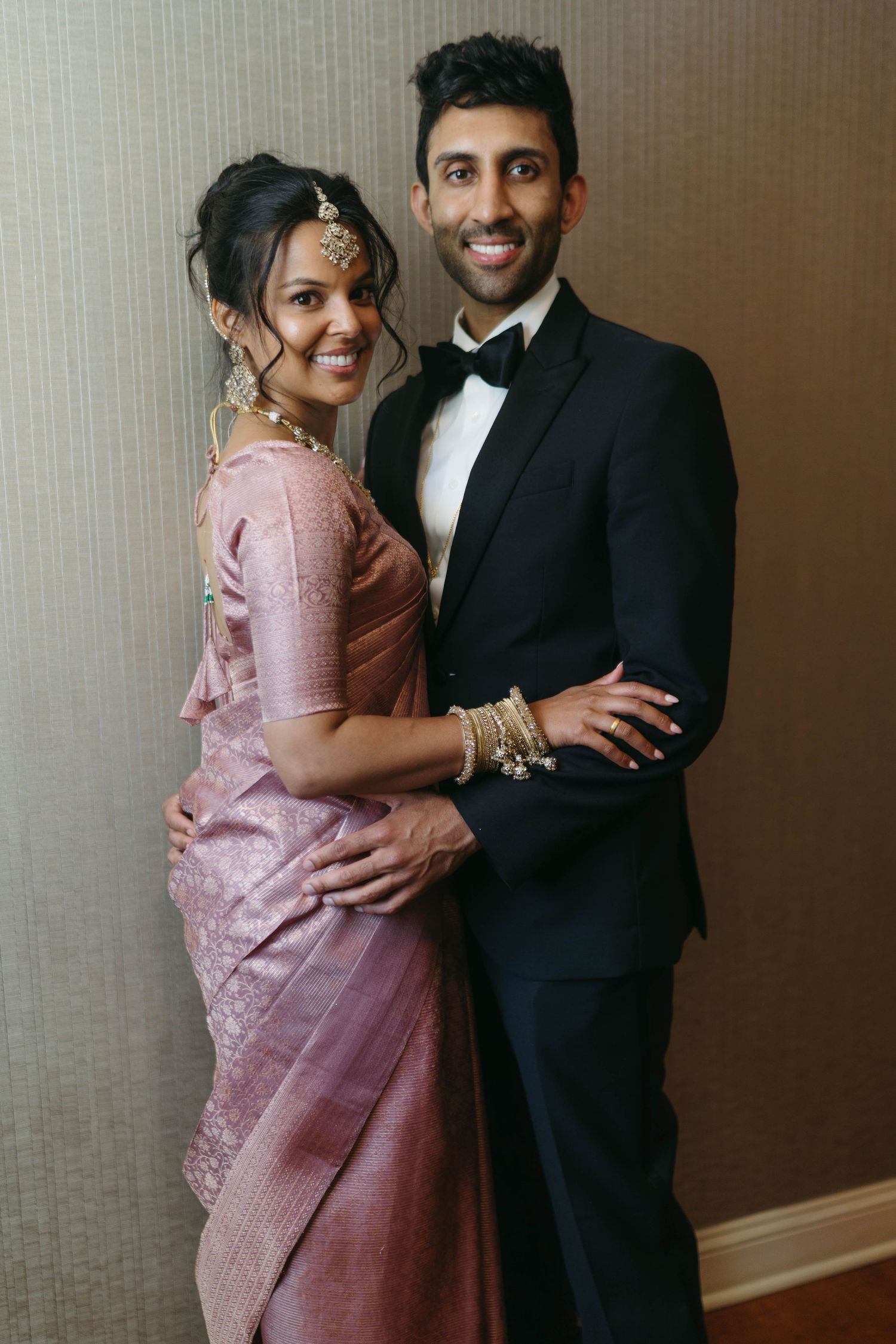 Indian bride in pink silk saree with gold jewelry posing with groom in black tuxedo.