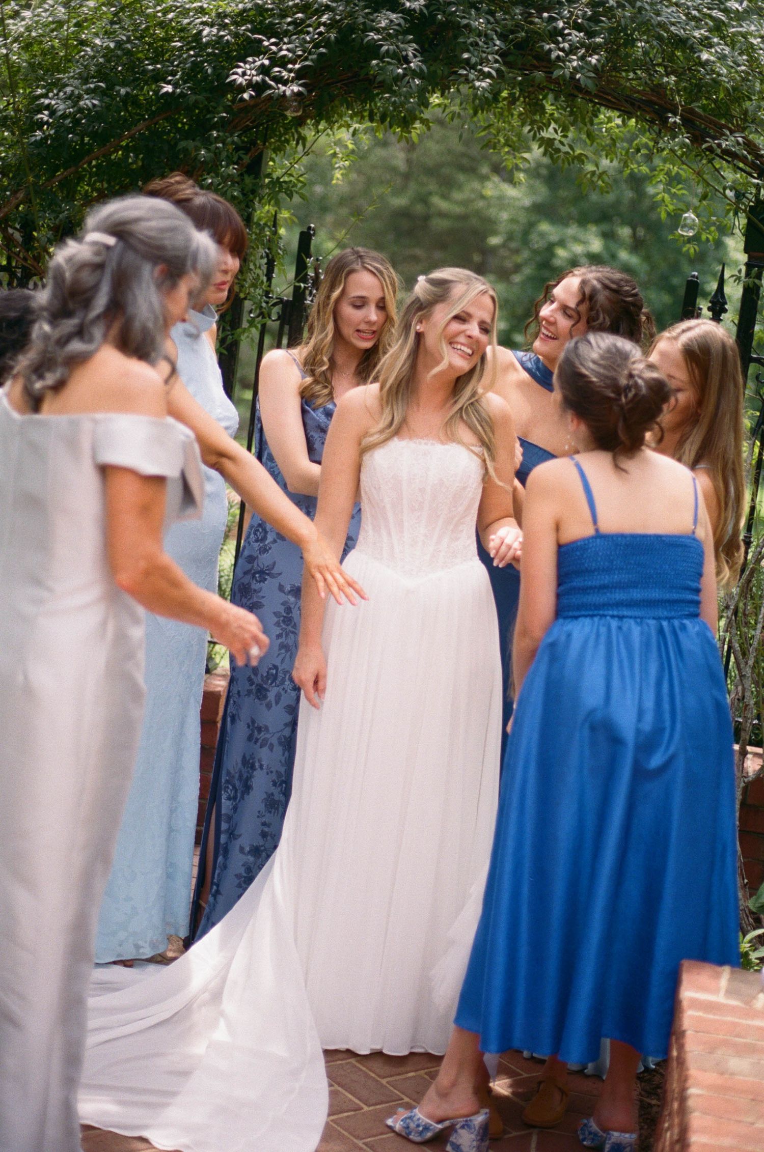 Bride in white corset gown smiles with bridesmaids in blue dresses under garden arbor.