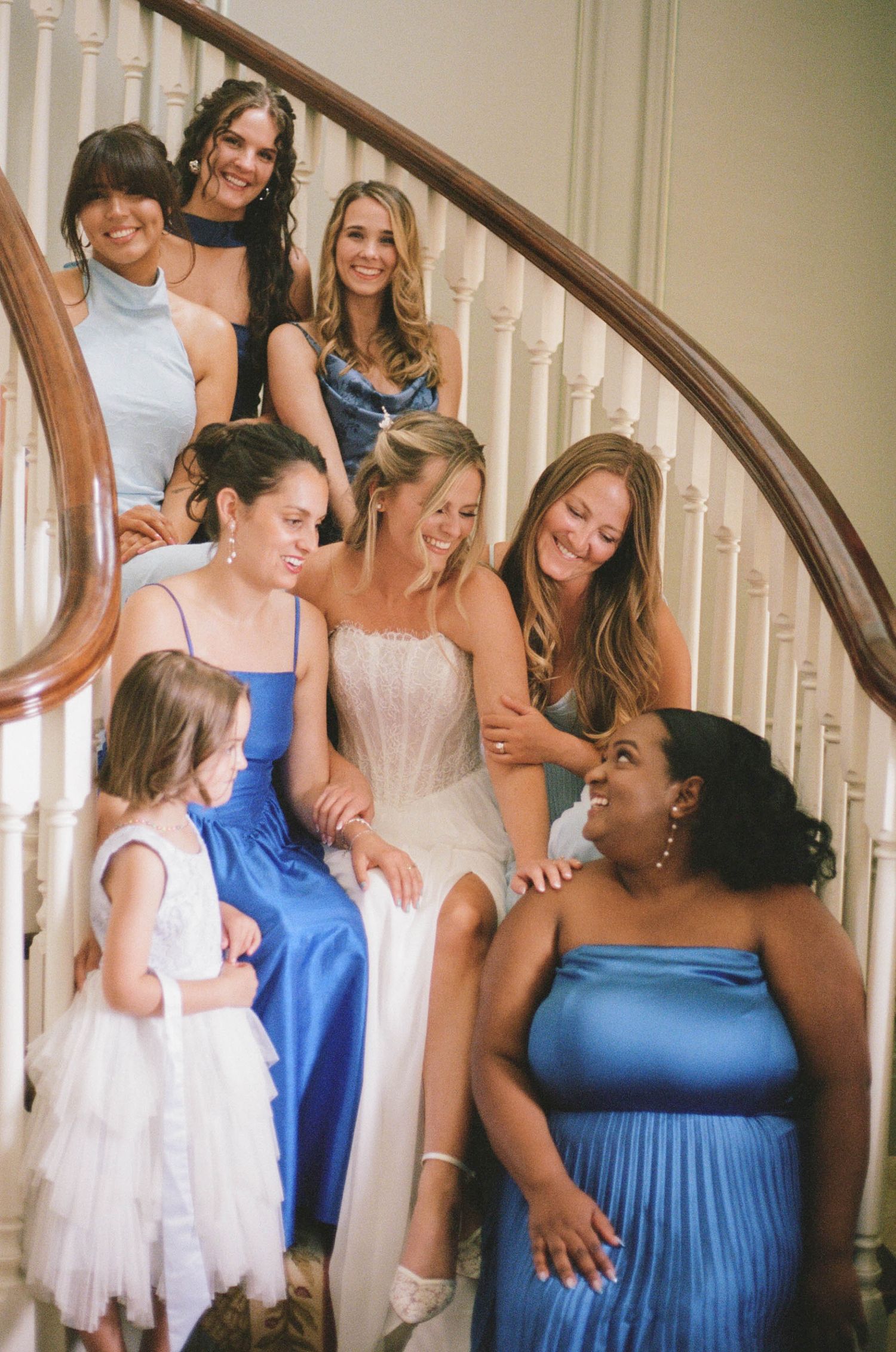 Bride surrounded by bridesmaids in blue dresses and a flower girl, laughing together on an elegant staircase.