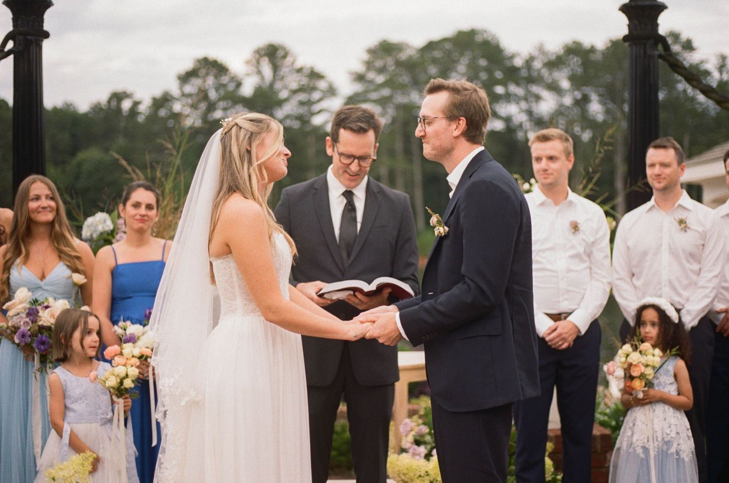 Couple exchanging vows at outdoor wedding ceremony with officiant, bridal party in blue dresses, and guests in background.