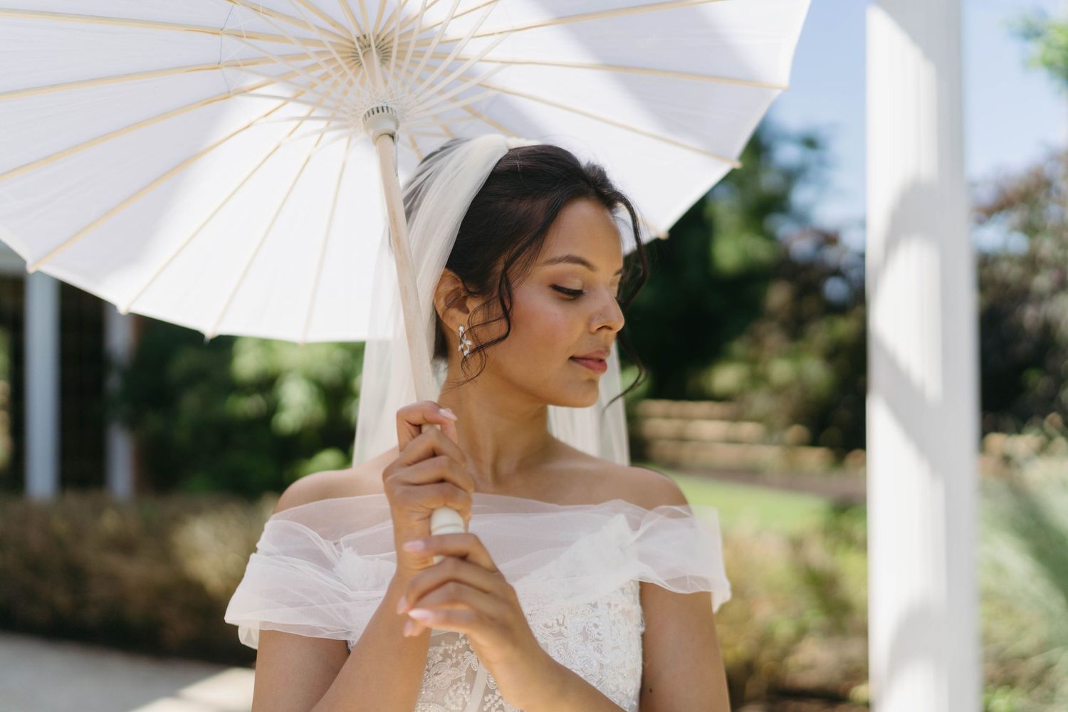 Elegant bride in off-shoulder gown holding white parasol umbrella outdoors on sunny wedding day.