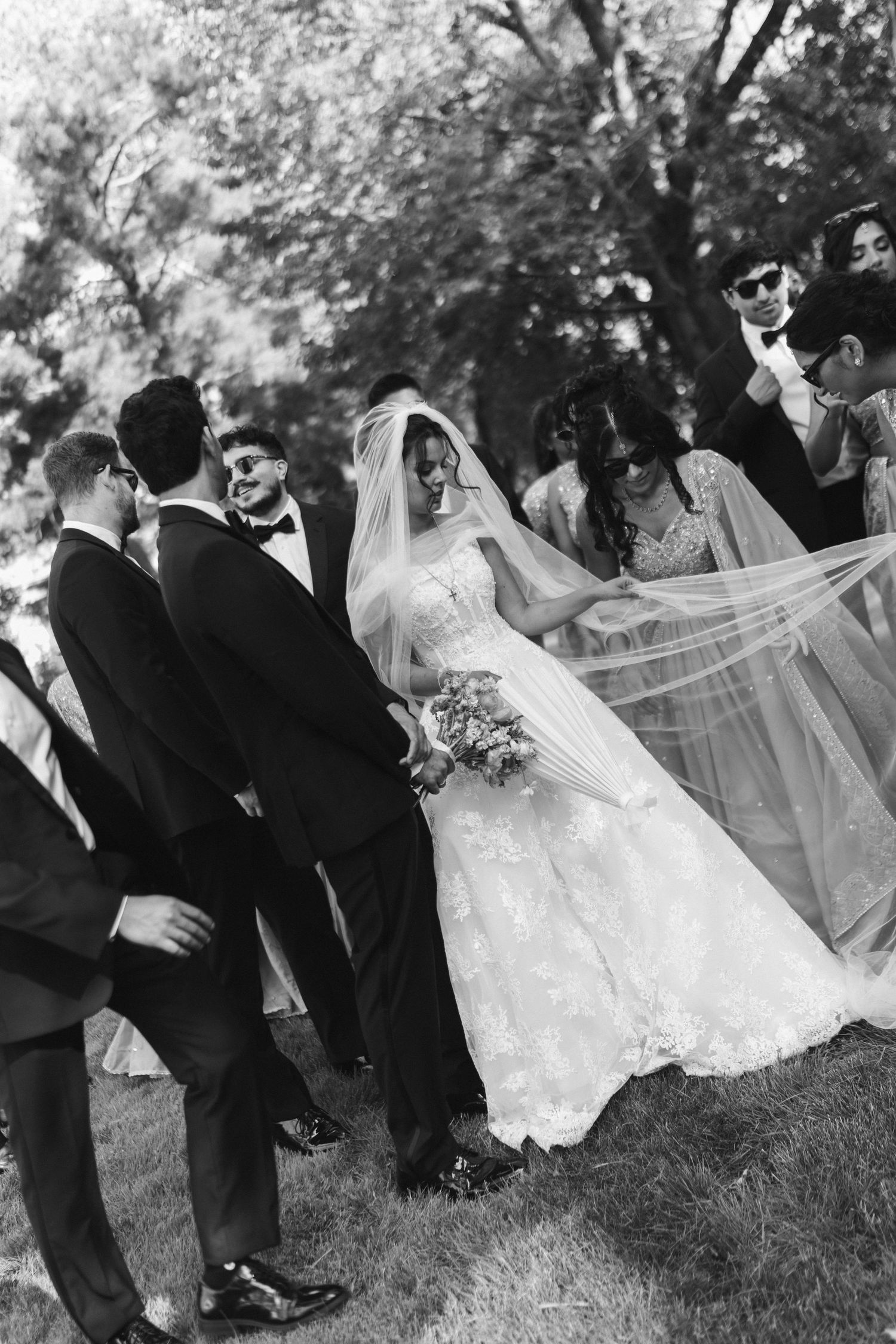 Black and white photo of bride and groom sharing a moment with wedding party outdoors among trees.