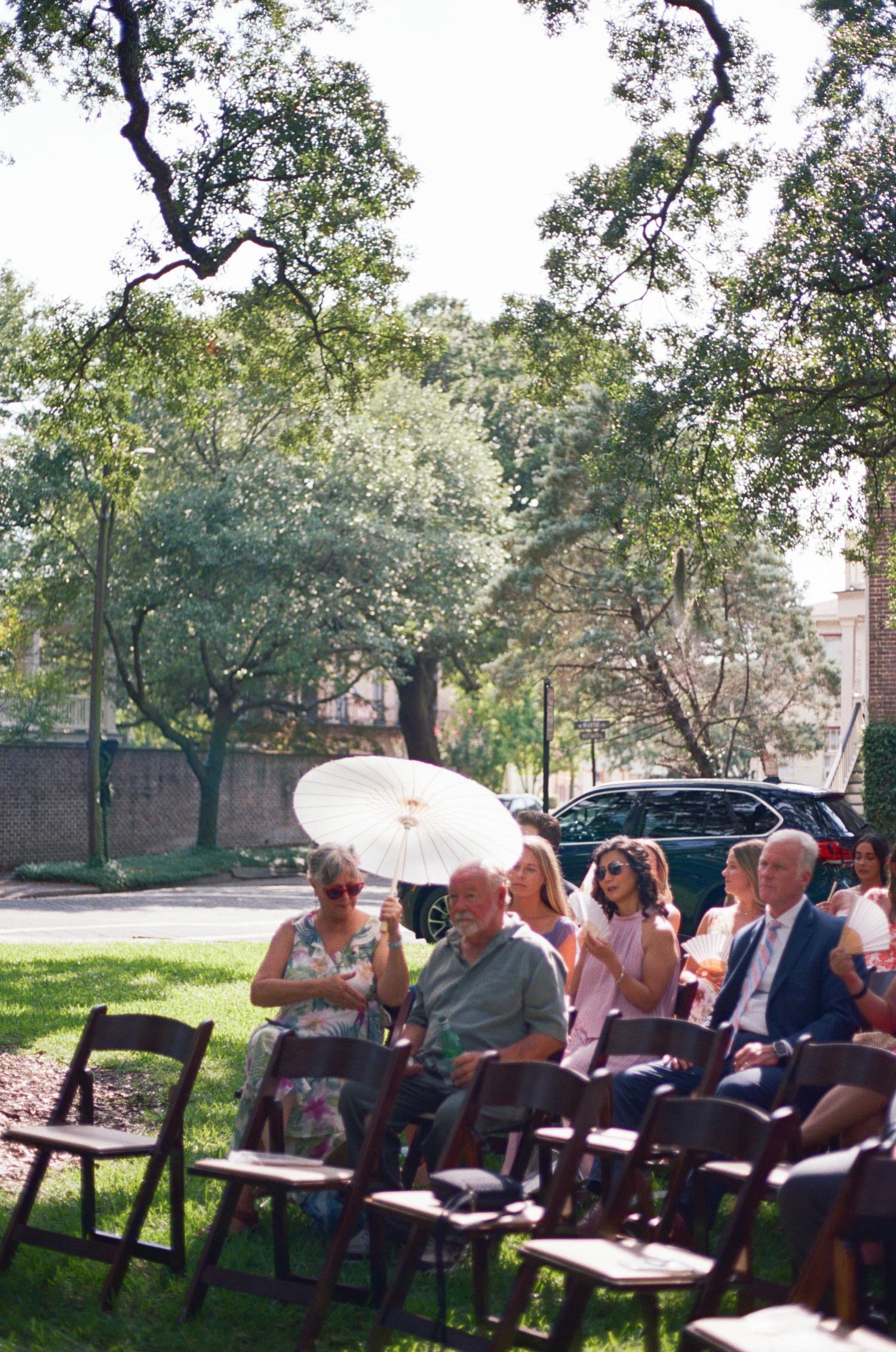 Wedding guests seated on folding chairs under oak trees with a white parasol visible.