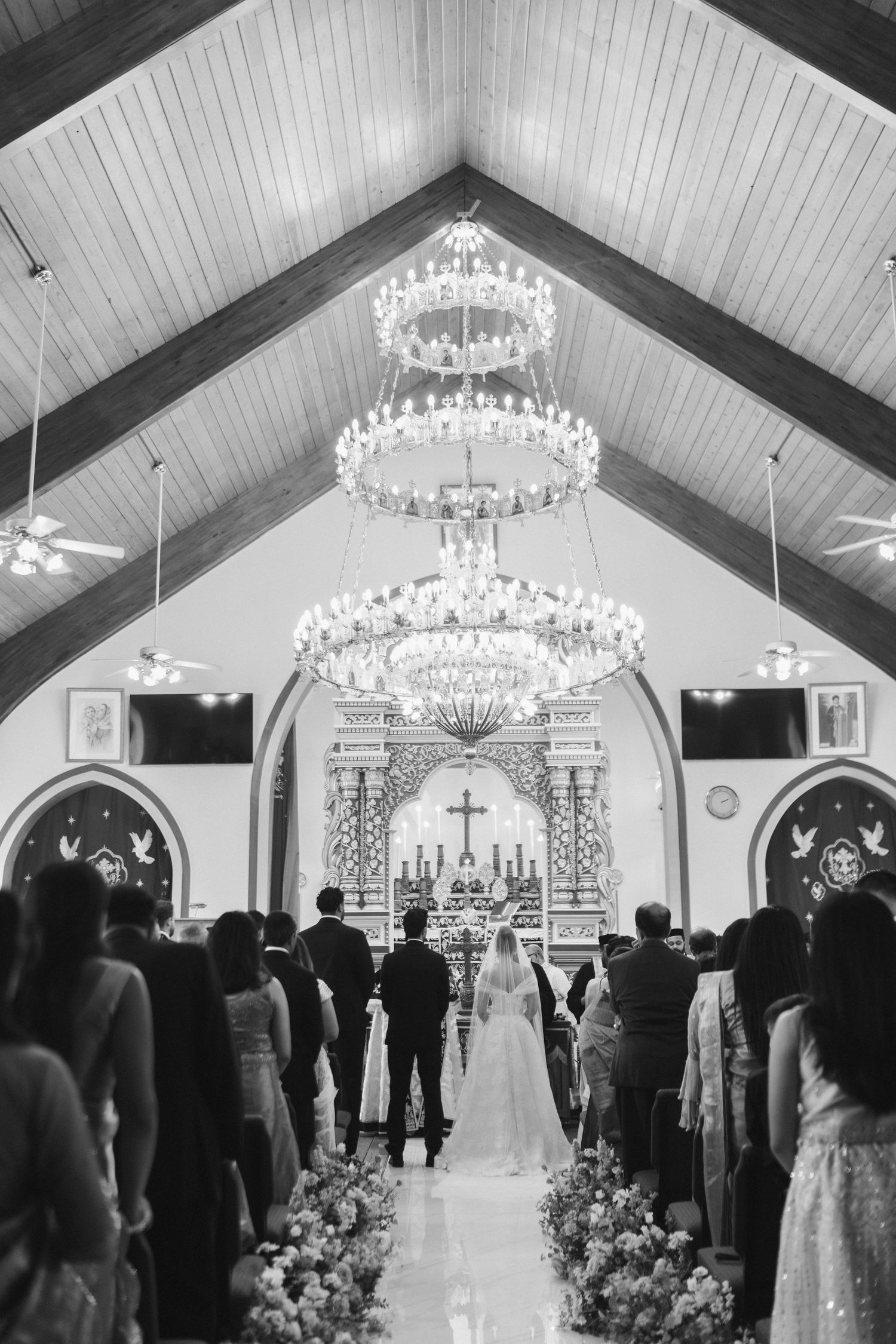 Black and white photo of a wedding ceremony inside a church with crystal chandeliers and floral aisle.