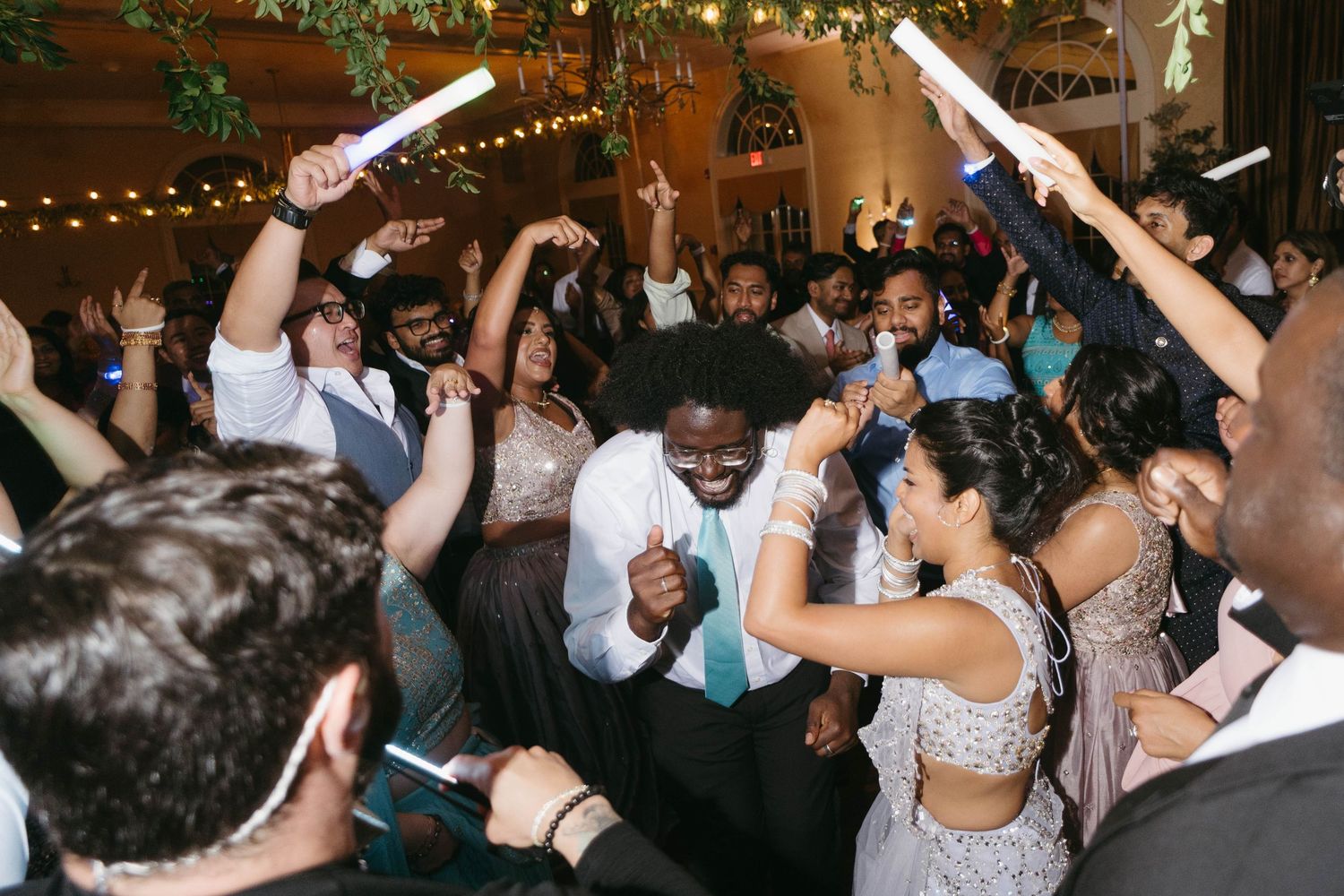Joyful wedding reception dance floor with guests celebrating, holding glow sticks, dressed in formal attire under warm string lights.