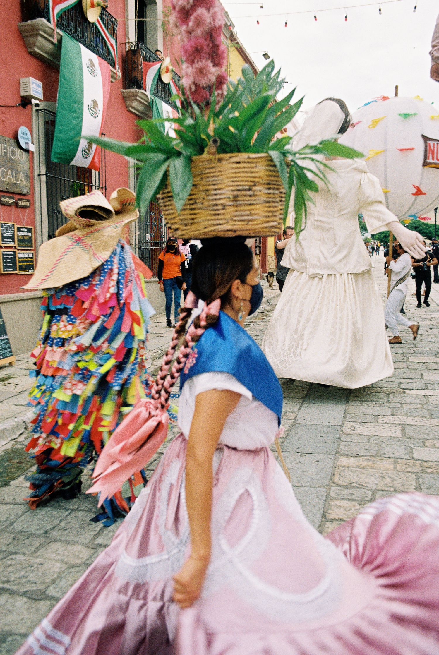 Wedding procession with dancers in pink dresses and colorful decorative elements on a cobblestone street.