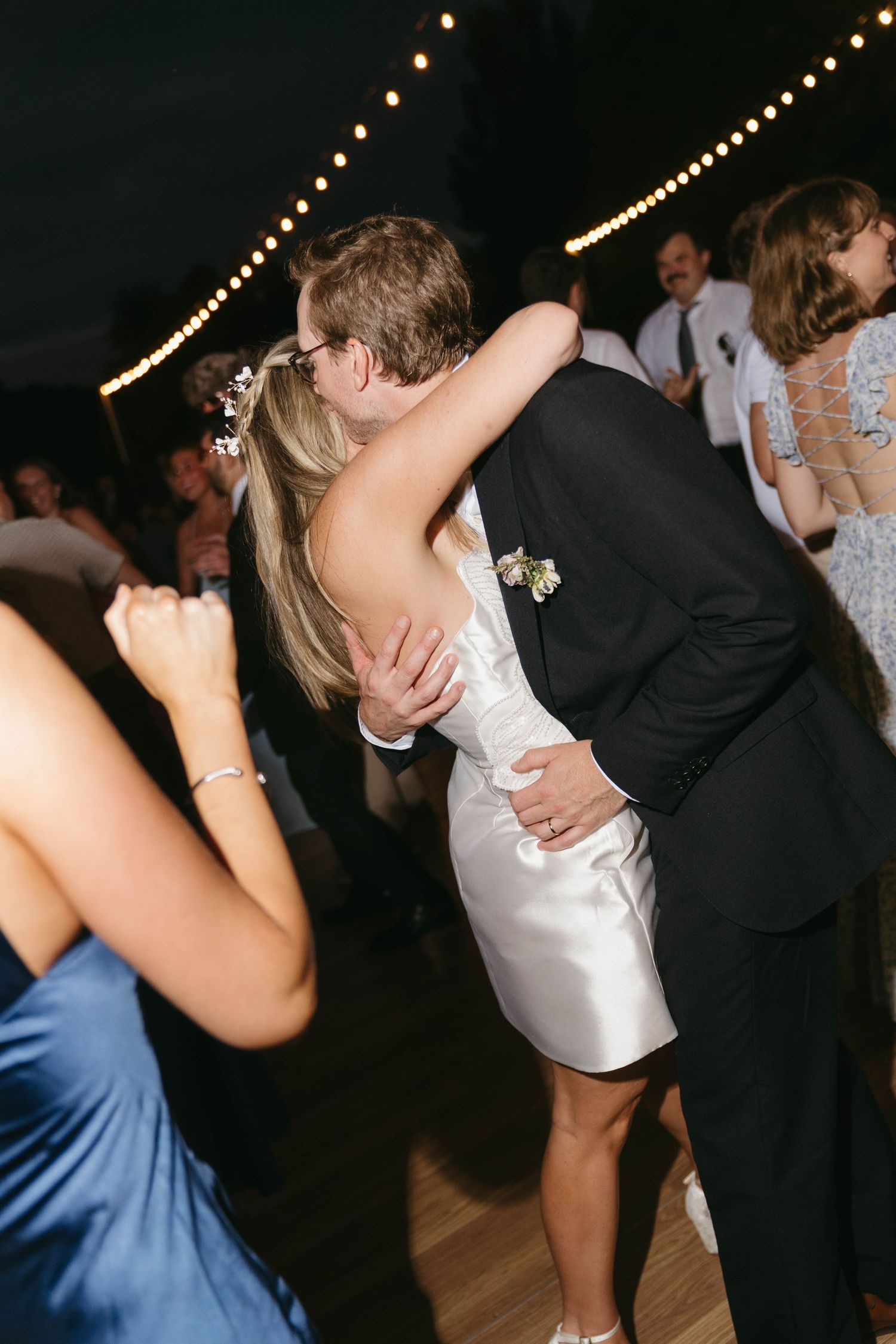 Bride and groom share a romantic kiss on the dance floor during their wedding reception under string lights.