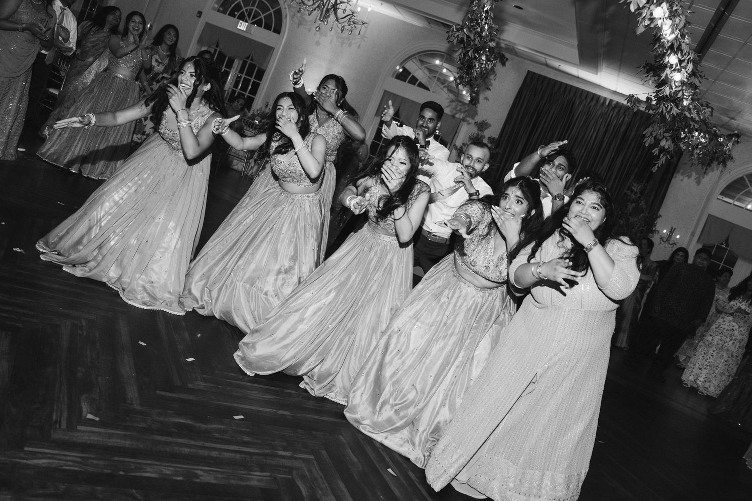 Bridesmaids in elegant ball gowns laughing and dancing together at a wedding reception in black and white photo.