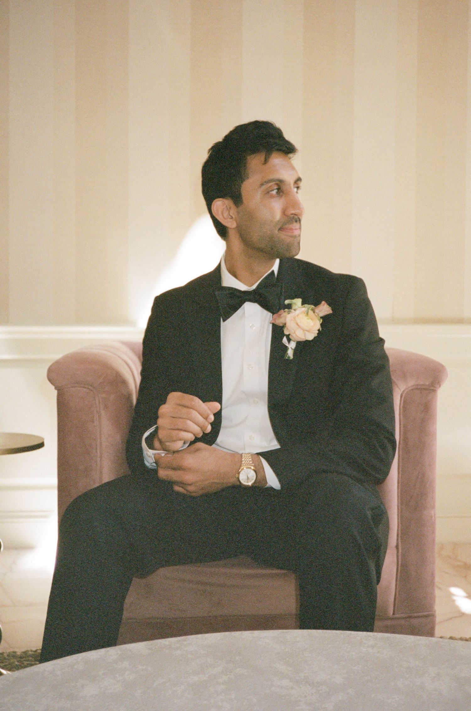 Dapper groom in black tuxedo with floral boutonniere sits relaxed in pink velvet chair at wedding.