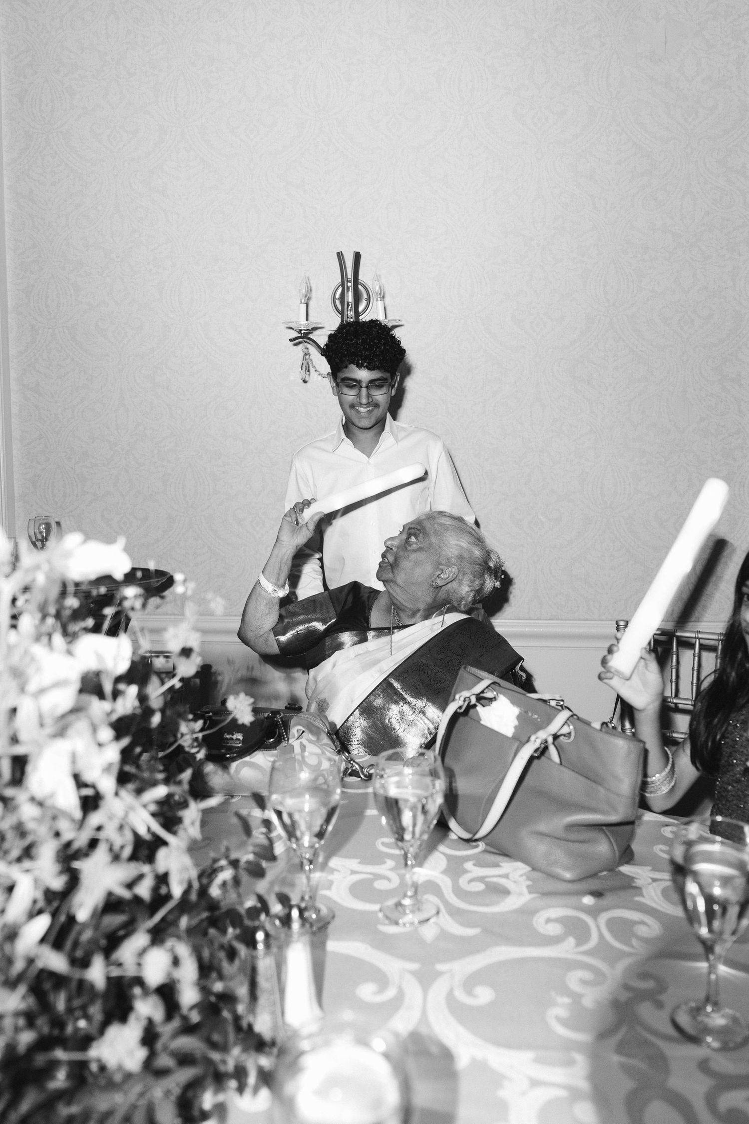 Black and white photo of guests laughing at a wedding reception dinner table with floral centerpieces.