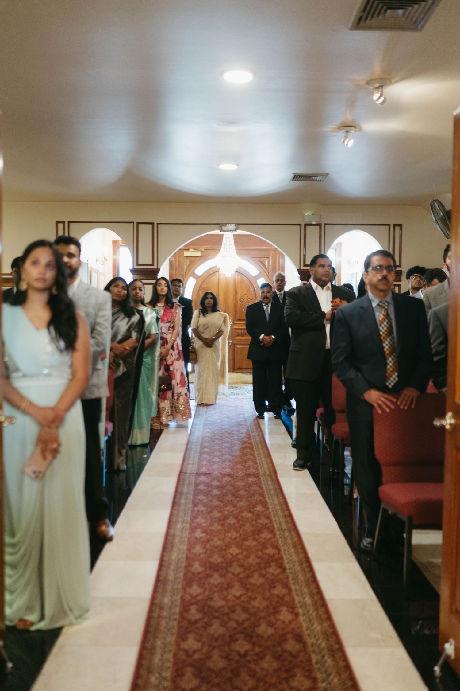 Guests stand in church pews along a decorated aisle runner during an Indian wedding ceremony.