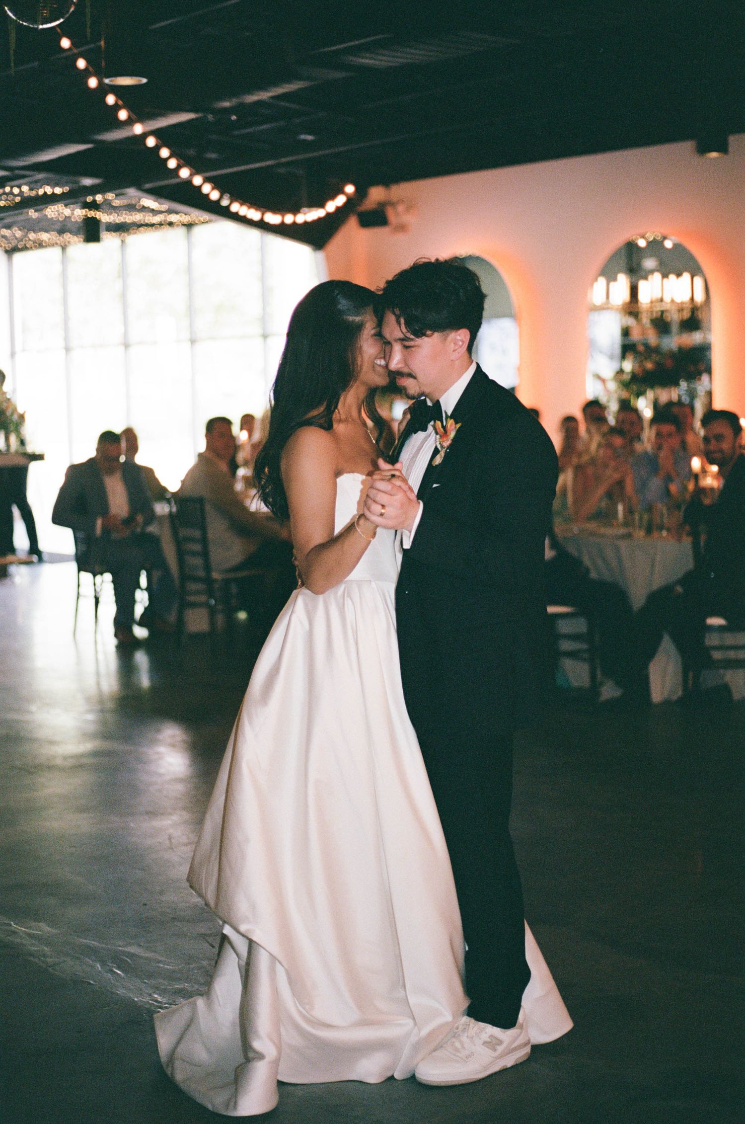Couples dance at an indoor wedding reception with string lights overhead.