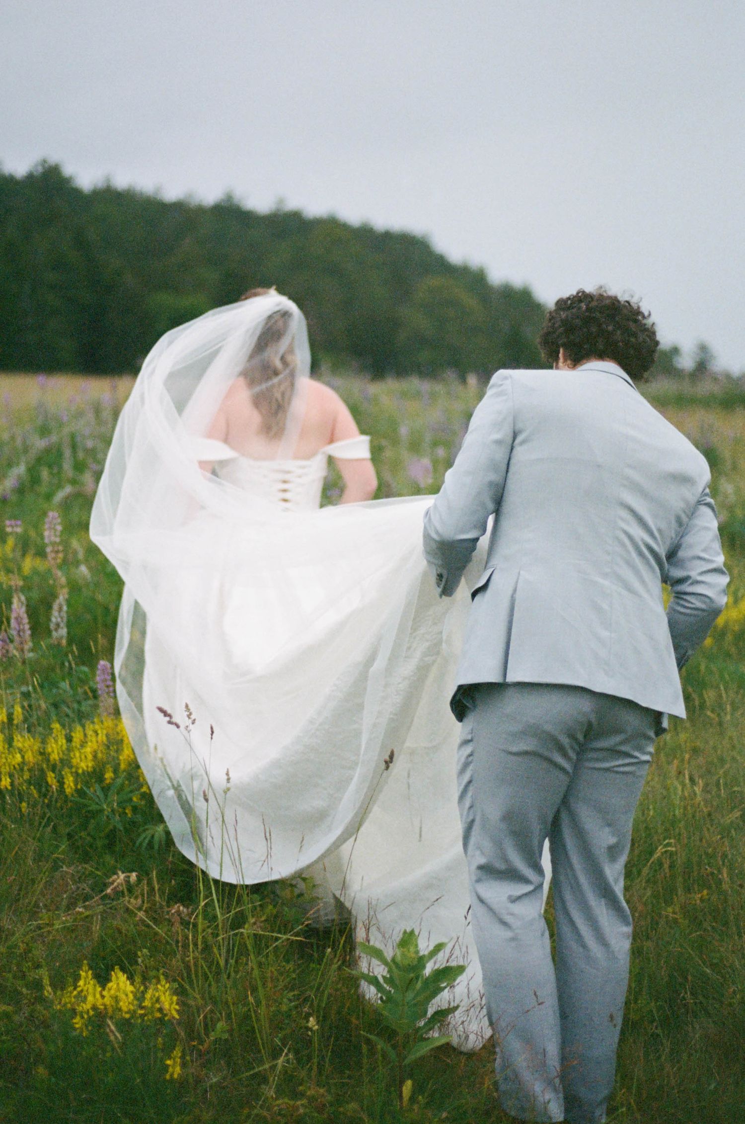 The train of a white wedding dress flows in the wind as couple walks through a meadow.
