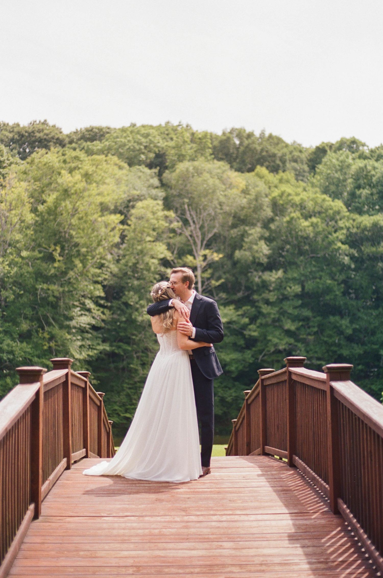 Bride and groom share a romantic kiss on a wooden bridge surrounded by lush green trees on their wedding day.