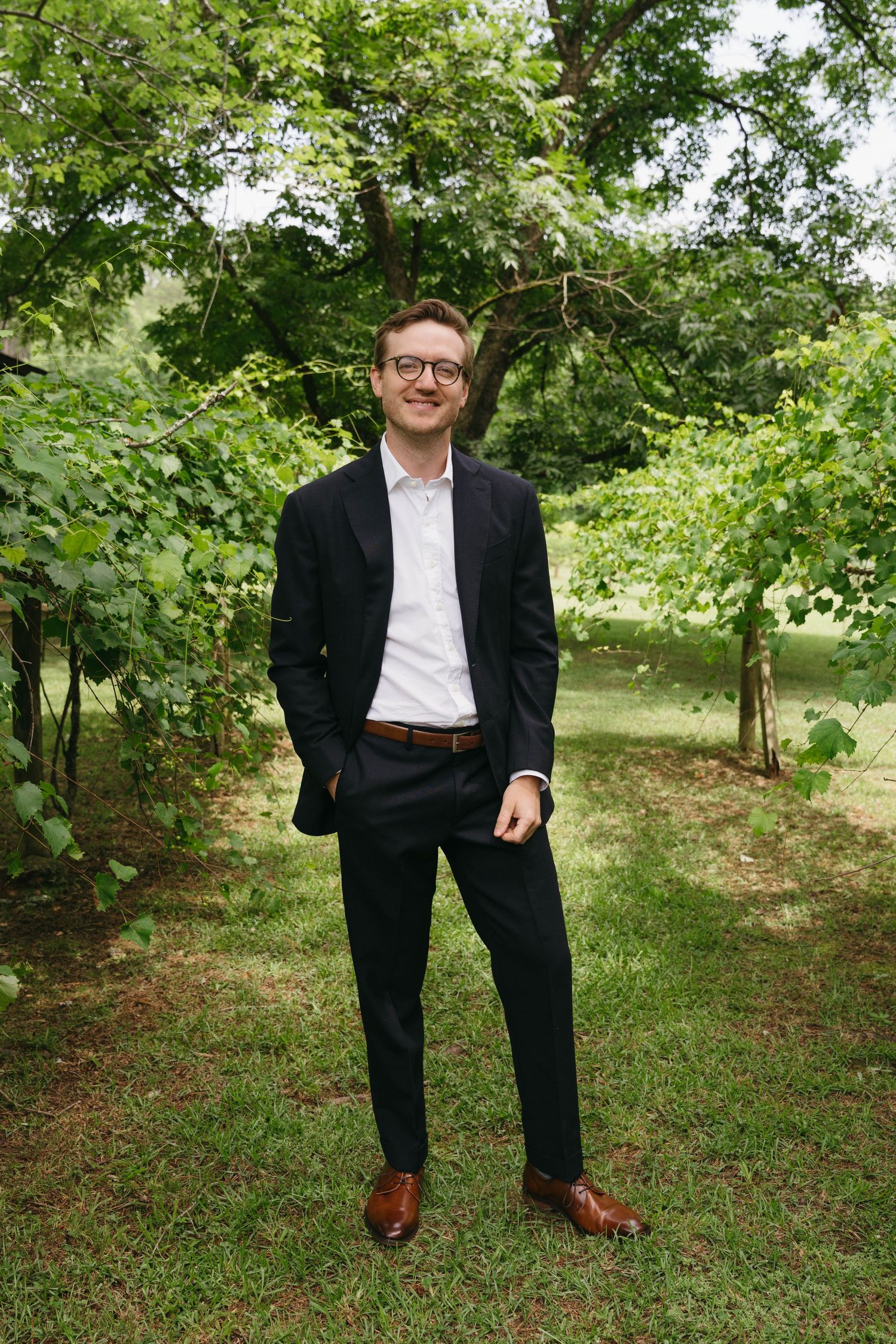 Smiling groom in black suit and glasses standing outdoors among lush green trees.