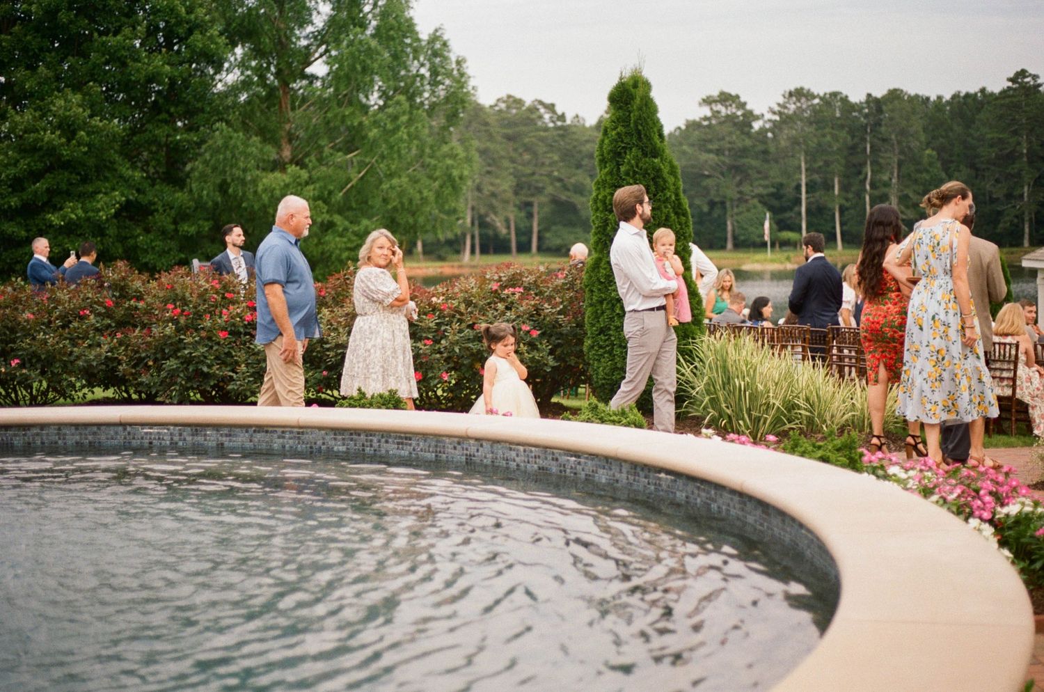 Guests mingle around an elegant outdoor fountain at a garden wedding reception, surrounded by lush greenery and colorful flowers.