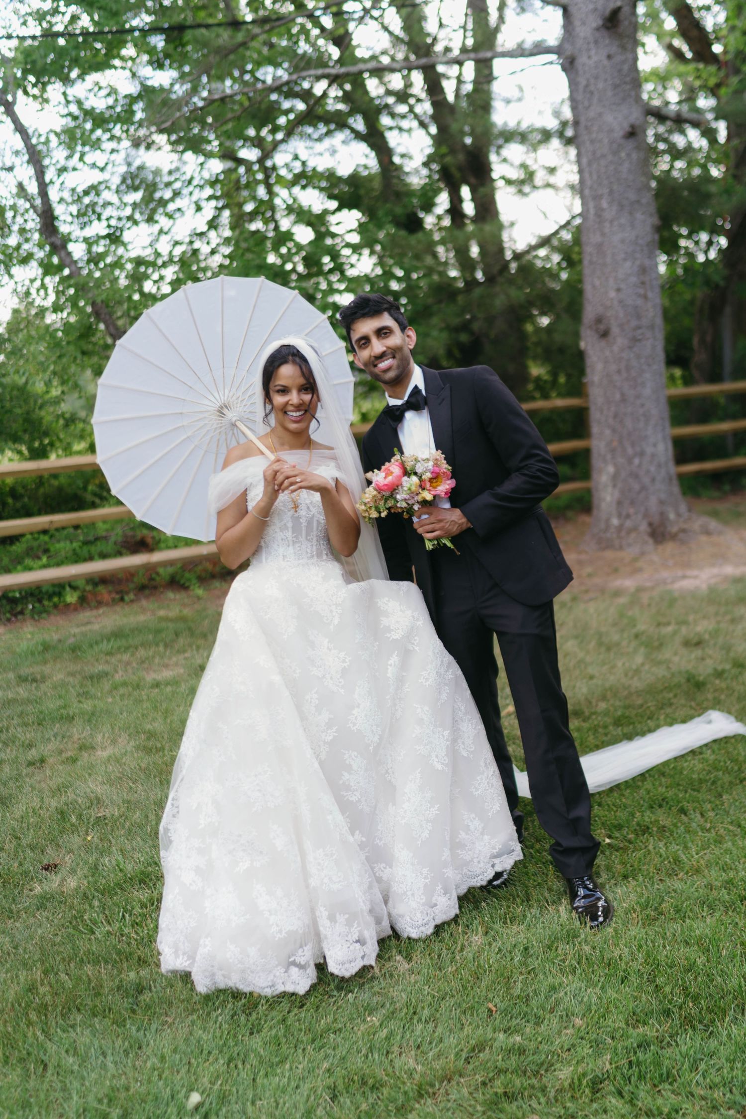 Smiling bride and groom posing outdoors with white parasol, bride in lace ballgown holding pink bouquet.
