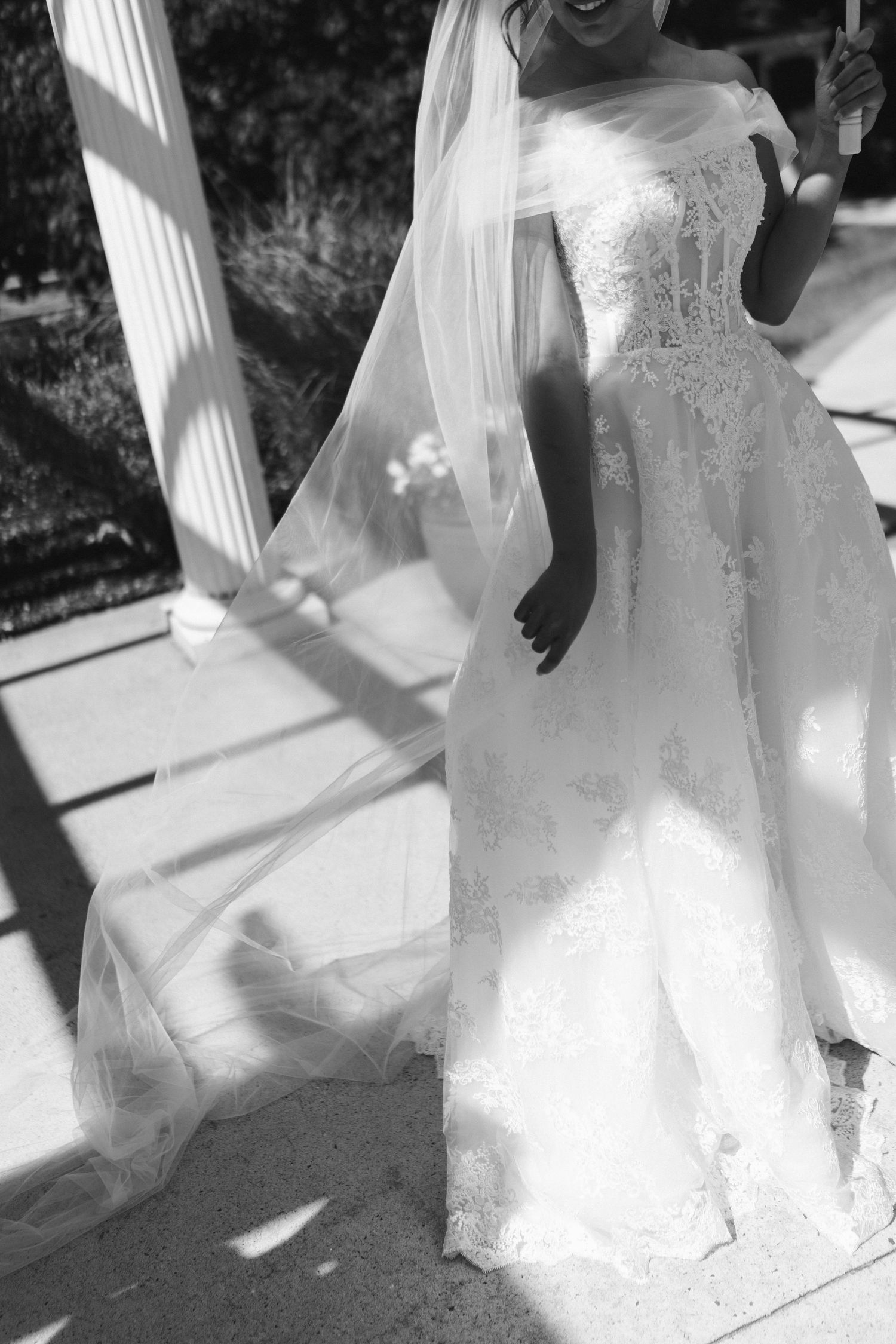 Black and white photo of bride in off-shoulder lace wedding gown with flowing veil outdoors.