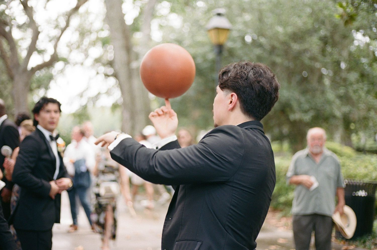 Outdoor wedding celebration with guests playing with orange balloons under trees.