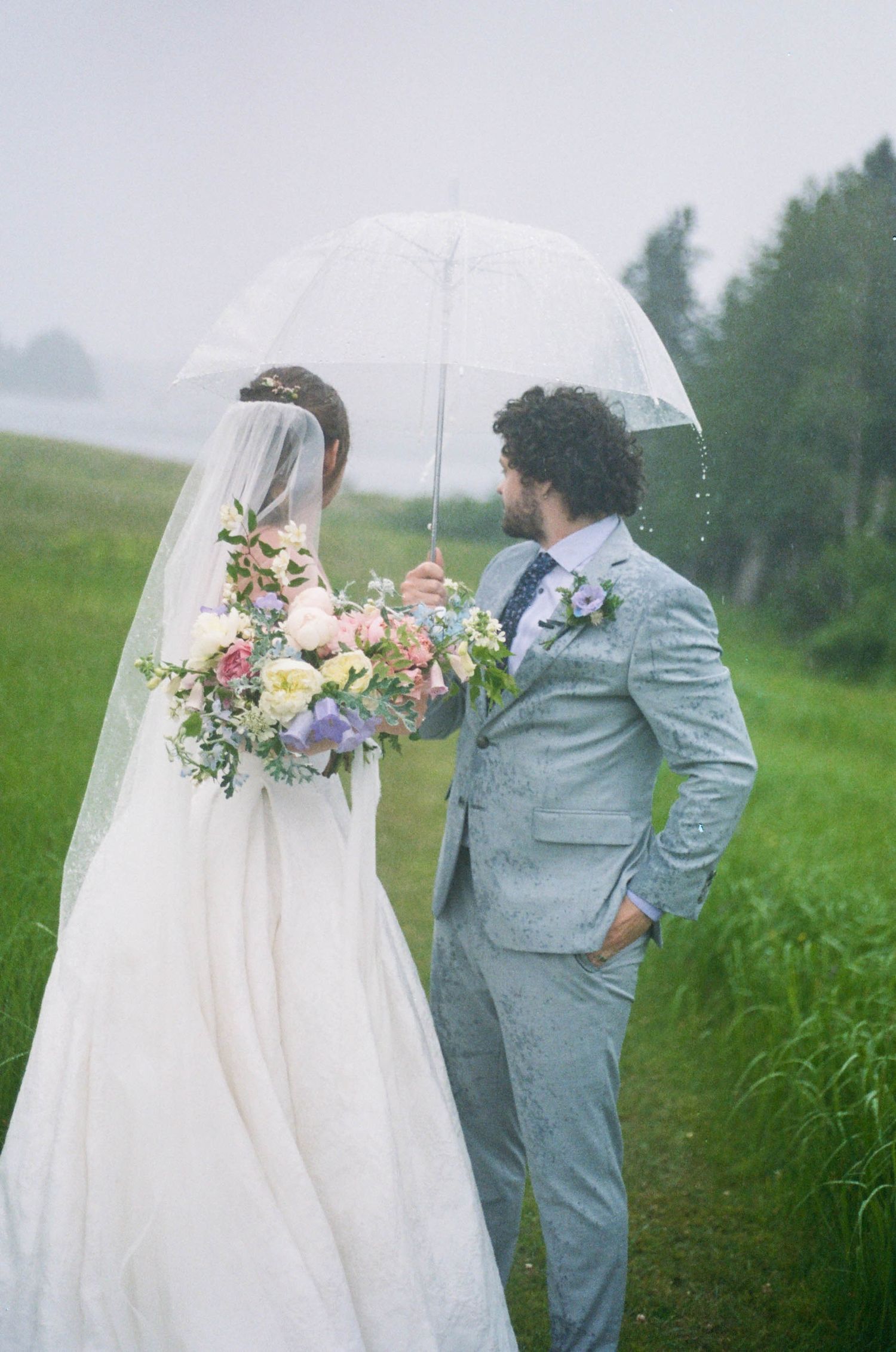 Wedding couple shares intimate moments under umbrellas in a lush green field.