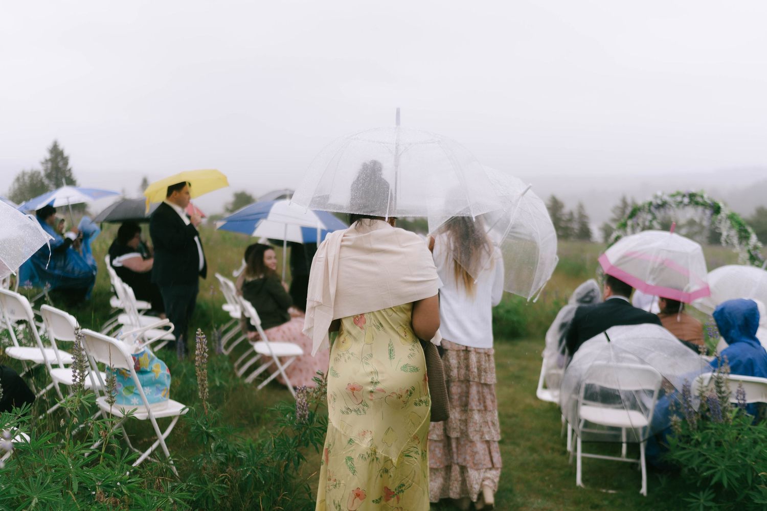 Wedding guests with umbrellas seated in white chairs during an outdoor ceremony.