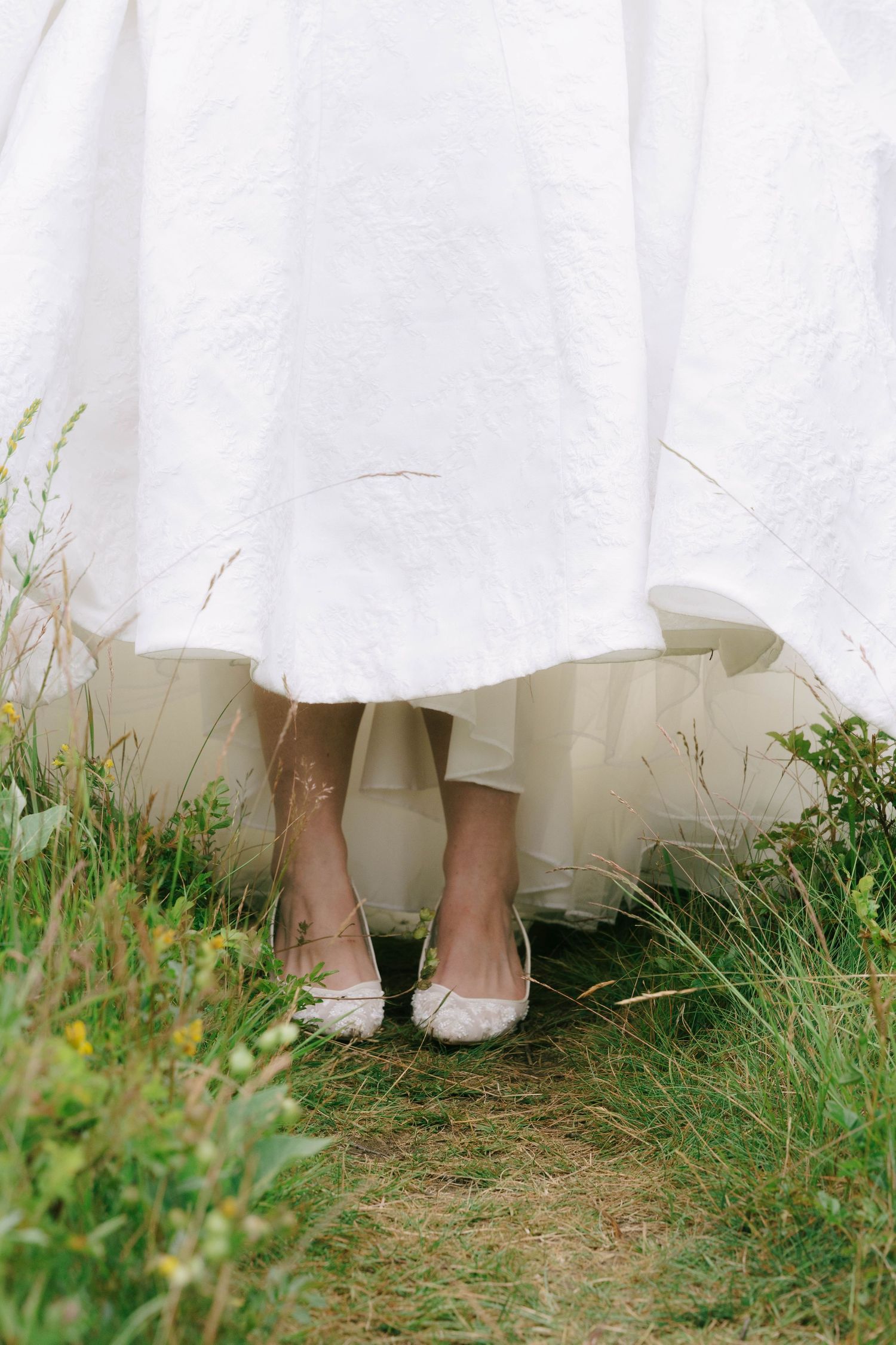 White wedding dress hem and flat bridal shoes peek out among green grass.