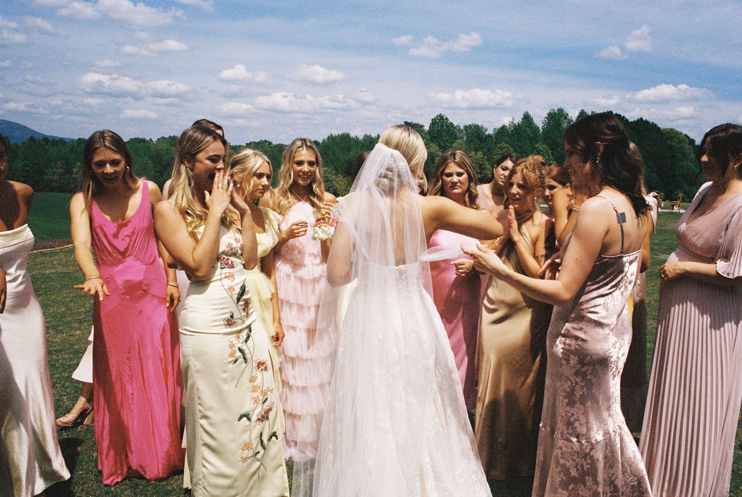Wedding guests in pastel dresses gather outdoors under bright summer skies.